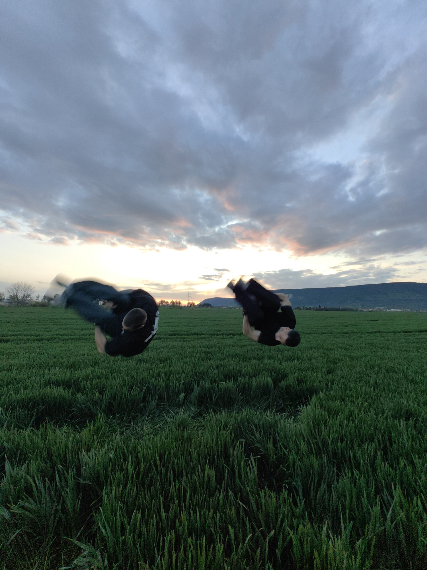 Two people are performing flips in a grassy field near Shumen, Bulgaria. The field is green with tall blades of grass. In the background, there is a sunset with cloudy skies. There are trees and a hill in the distance.