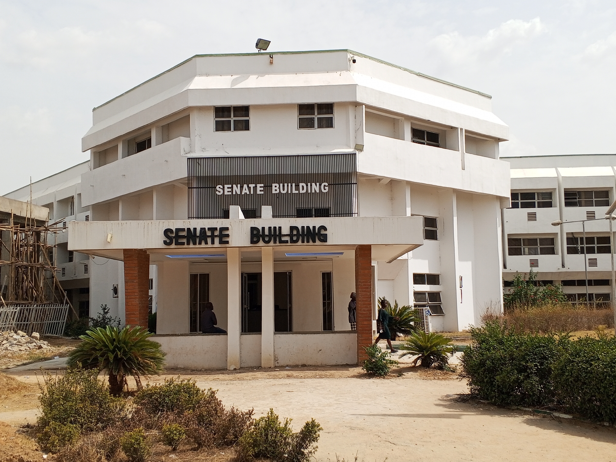The image shows the Senate Building at the Federal University of Technology, Minna, Nigeria. The building is a multi-story, white structure with a prominent sign reading "SENATE BUILDING" on both the upper and lower sections of the front façade. The building features a flat roof, rectangular windows, and a colonnaded entrance. The foreground includes dirt patches, sparse greenery, and small palm-like plants. There are a few people visible near the building's entrance. A second building is visible to the right, and there is construction with scaffolding on the left. The sky is overcast.