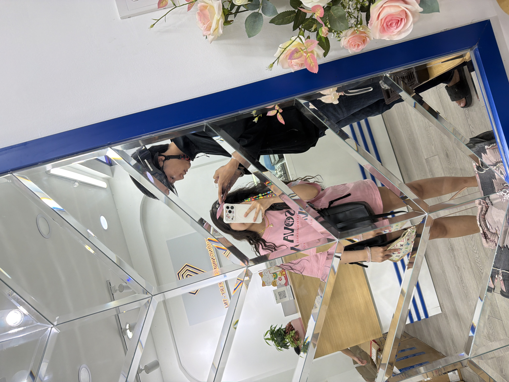 The provided image is oriented on its side, showcasing a complex reflection within a large, multi-faceted mirror display. The top portion of the image features a white ceiling area with a decorative arrangement of artificial pink and white roses and green leaves, positioned above a horizontal blue border that frames the top of the mirror.
Reflected prominently in the mirror's various panels is a woman taking a selfie with a white smartphone. She has long dark hair and is wearing a pink t-shirt with the reversed text "AVOCAT" (or similar) visible on it, along with a black shoulder bag. A colorful beaded strap adorns her wrist, connected to her phone. In her other hand, she holds a smaller handbag, which appears to feature a monogram pattern resembling Louis Vuitton.
Partially visible next to her reflection is a man wearing a black shirt and a black helmet, possibly a motorcycle helmet, with the brand "ROCKBROS" faintly legible on its side. His face is seen in profile.
The geometric mirror panels create multiple, fragmented reflections of the interior space. These reflections include a wooden-patterned floor, various light fixtures, a small potted green plant on a shelf, and a QR code. Reversed text, possibly "CATT-HAT," is also discernible on a reflected sign. The scene depicts an indoor setting, likely a shop or cafe in Da Nang, Vietnam, where the unique mirror installation serves as a photo opportunity. The environment is well-lit, suggesting daytime.
