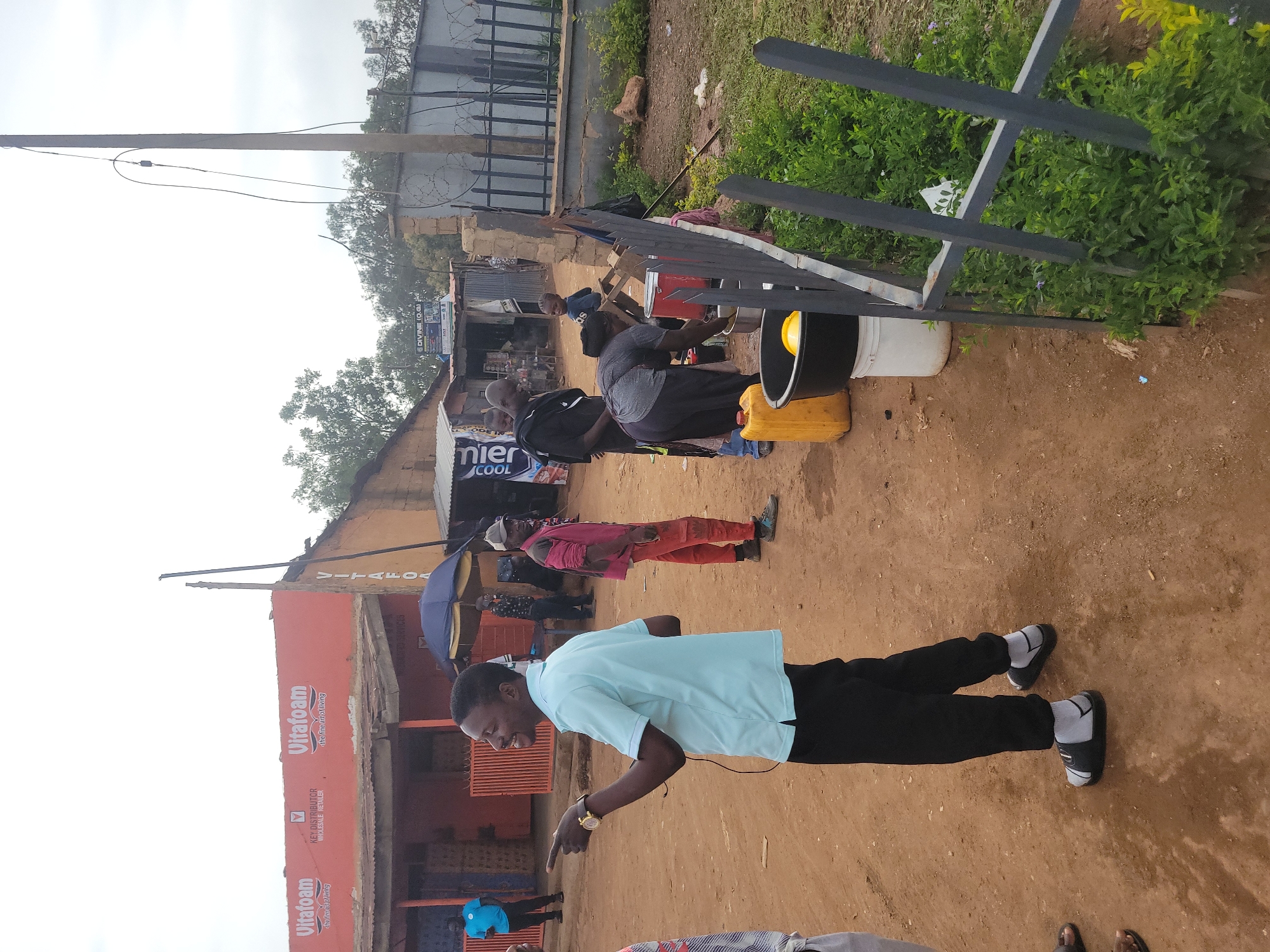 In Gura Top, Nigeria, a group of people are gathered in front of a building with "Vitafoam" signage. A man in a light blue polo shirt and black pants, wearing sandals and socks, is pointing his left hand outwards, appearing to speak. Beside him, a person in a red shirt and orange pants stands. To the right, a woman in a grey shirt and black pants is washing items in a large basin. A yellow jerrycan and a white bucket are visible nearby. A black umbrella is open over another person in the background. The ground is dirt, and a metal fence with overgrown green bushes is present. In the background, there are more buildings and trees under a cloudy sky.