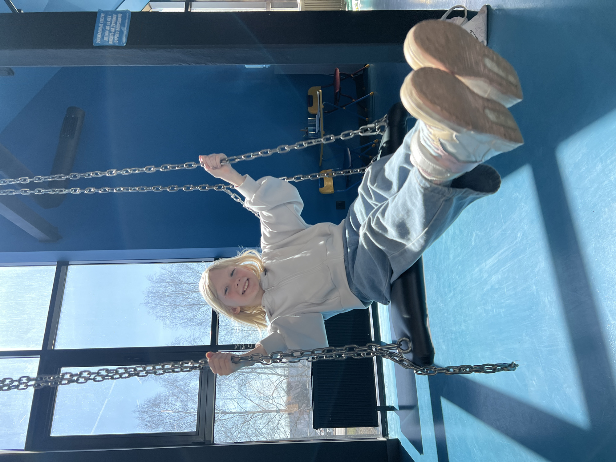 A young girl is playing on a chain swing inside a building in Izhevsk, Russia. She is holding onto the chains and her feet are raised in the air. The walls of the building are a dark blue. There are large windows in the background.