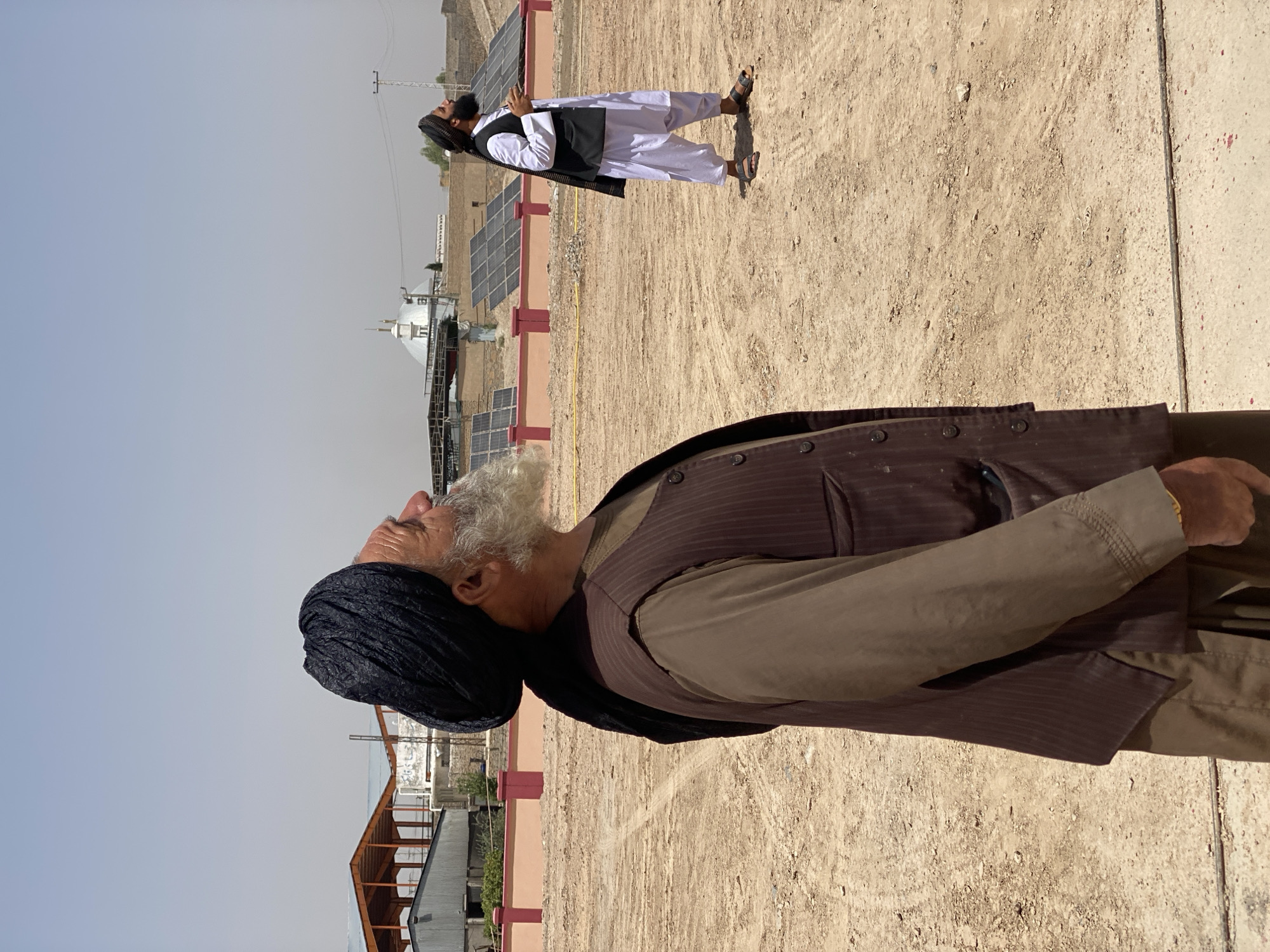 In an outdoor, dusty environment, two men stand in the daytime. The man in the foreground is an elderly individual with a long, grey beard and a black turban, looking upwards. He is wearing a brown long-sleeved shirt and a dark pinstriped vest. To the right, a younger man with a black turban and a beard is also looking upwards. He is dressed in a white shalwar kameez and a black vest. Behind them, several solar panels are arranged in rows against a low wall. In the background, there are buildings with a domed structure and what appears to be a covered market or industrial area. The ground is predominantly dirt and sand, with some scattered debris and a thin metal wire. The sky is clear and bright.