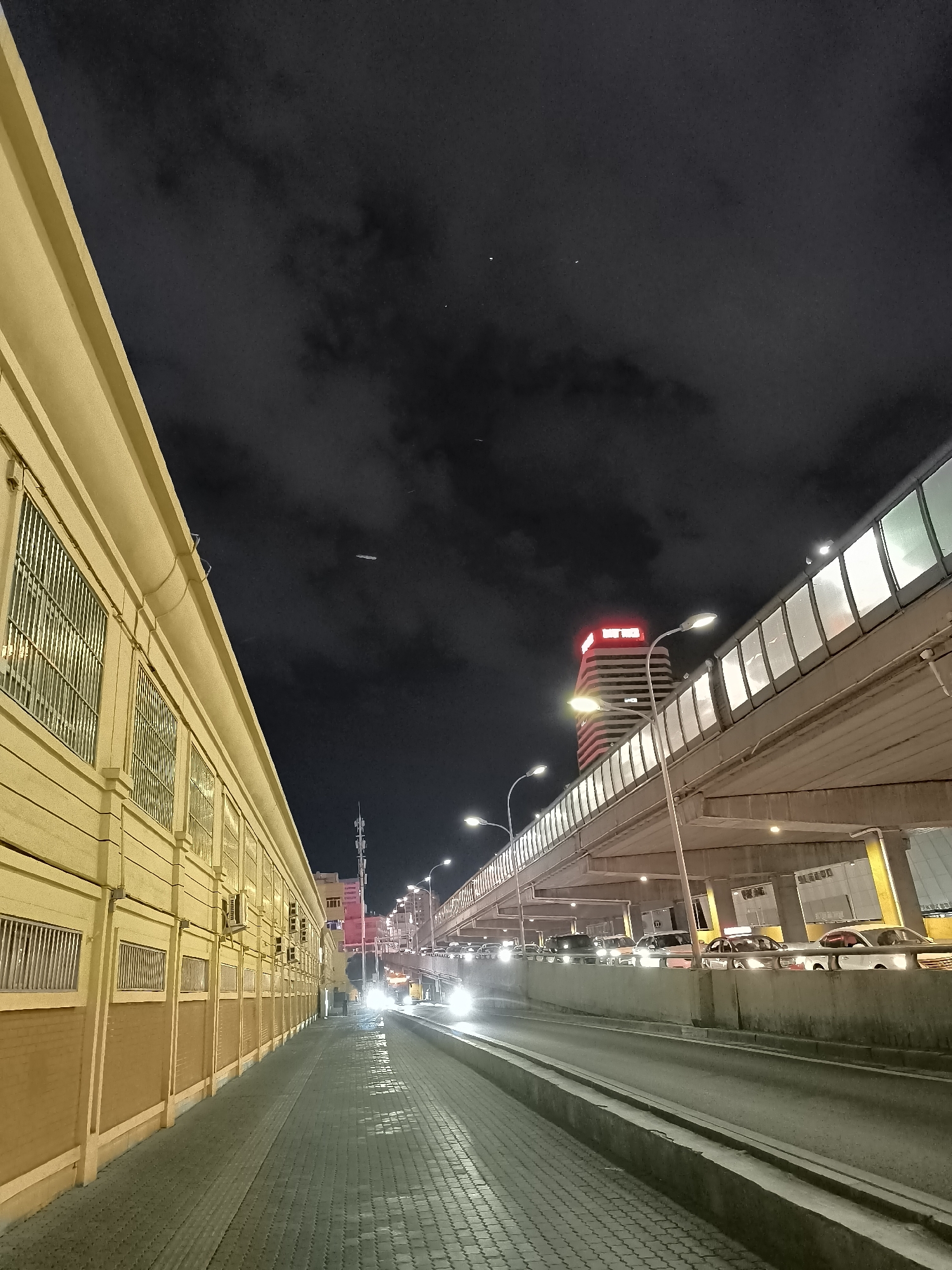The image depicts an elevated highway overpass at night in Ürümqi, China. A long, yellow building with barred windows runs along the left side of the frame, adjacent to a pedestrian walkway paved with interlocking stones. The overpass, illuminated by streetlights, carries traffic, with cars visible in both directions. The sky is dark and cloudy, with a few distant lights and a faint airplane contrail. In the background, a tall building with illuminated red signage stands out. The overall scene is characteristic of a busy urban environment at night, with the architecture and lighting suggesting a developed area.