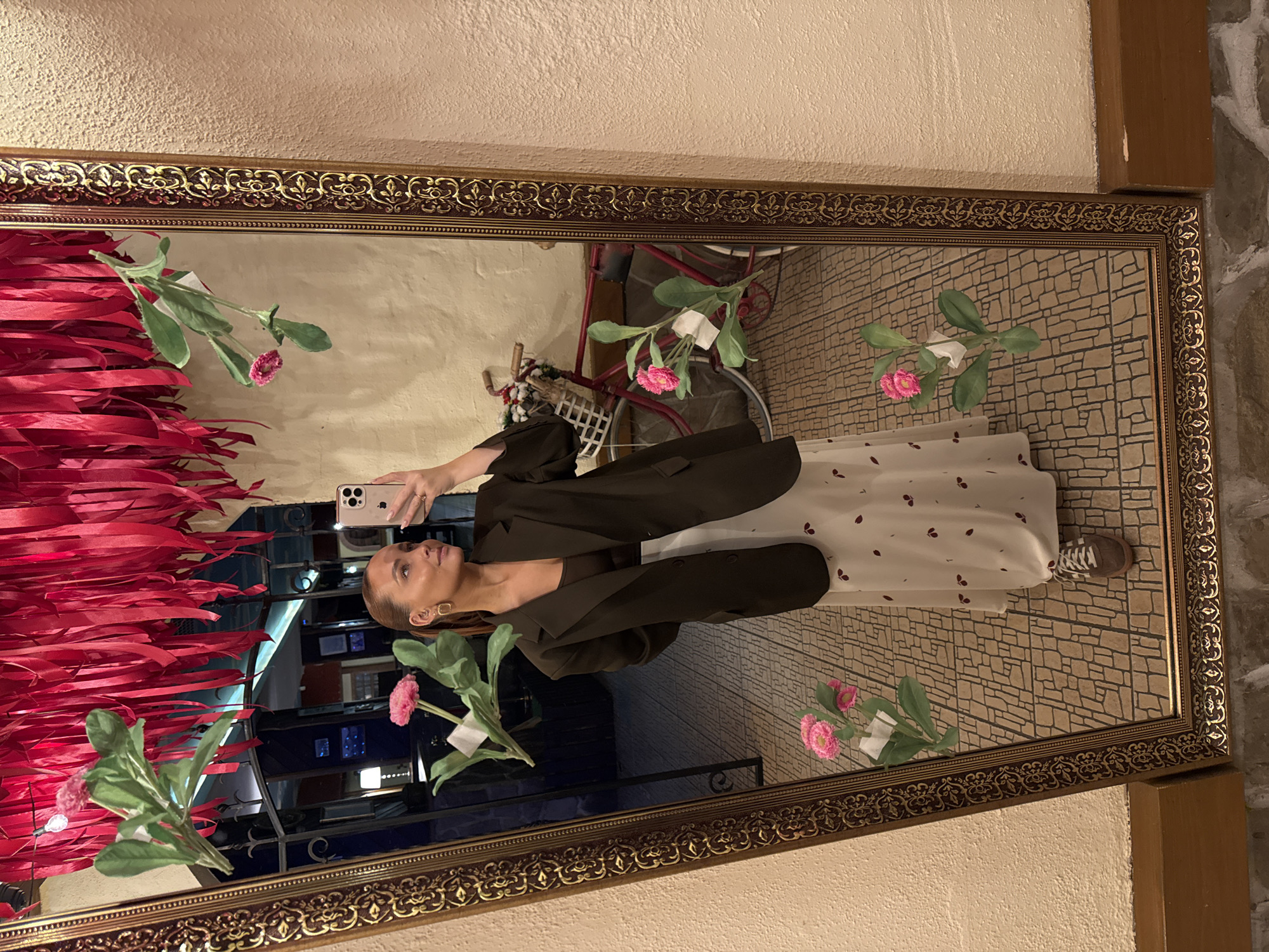 An indoor scene in Chaikovsky, Russia, depicts a young adult female taking a mirror selfie. The subject, positioned centrally, has light skin, brown hair pulled back, and wears a dark brown oversized blazer over a dark brown top, a light-colored long skirt with a scattered pattern of small dark red shapes, and brown sneakers with white laces. She holds a light-colored smartphone with a multi-lens camera array in her right hand, angled towards her reflection. Gold-toned hoop earrings are visible, along with a ring on her left ring finger.
The subject is reflected in a large, rectangular mirror featuring an ornate, dark gold-toned frame with intricate carvings. The reflected environment behind her includes a ceiling densely covered with numerous hanging red fabric ribbons. To her right, a light beige textured wall is visible. The reflected floor displays a geometric pattern of light brown and beige tiles. In the mid-ground of the reflection, a red vintage-style bicycle equipped with a white wicker basket on its handlebars, containing decorative items, is present. A darker, dimly lit interior space is visible further in the background. Several artificial pink flowers with green leaves are affixed directly to the mirror's surface, secured by small white adhesive pads. The wall to the right of the mirror's frame is a light beige textured surface, with a dark wooden baseboard visible along its lower edge.