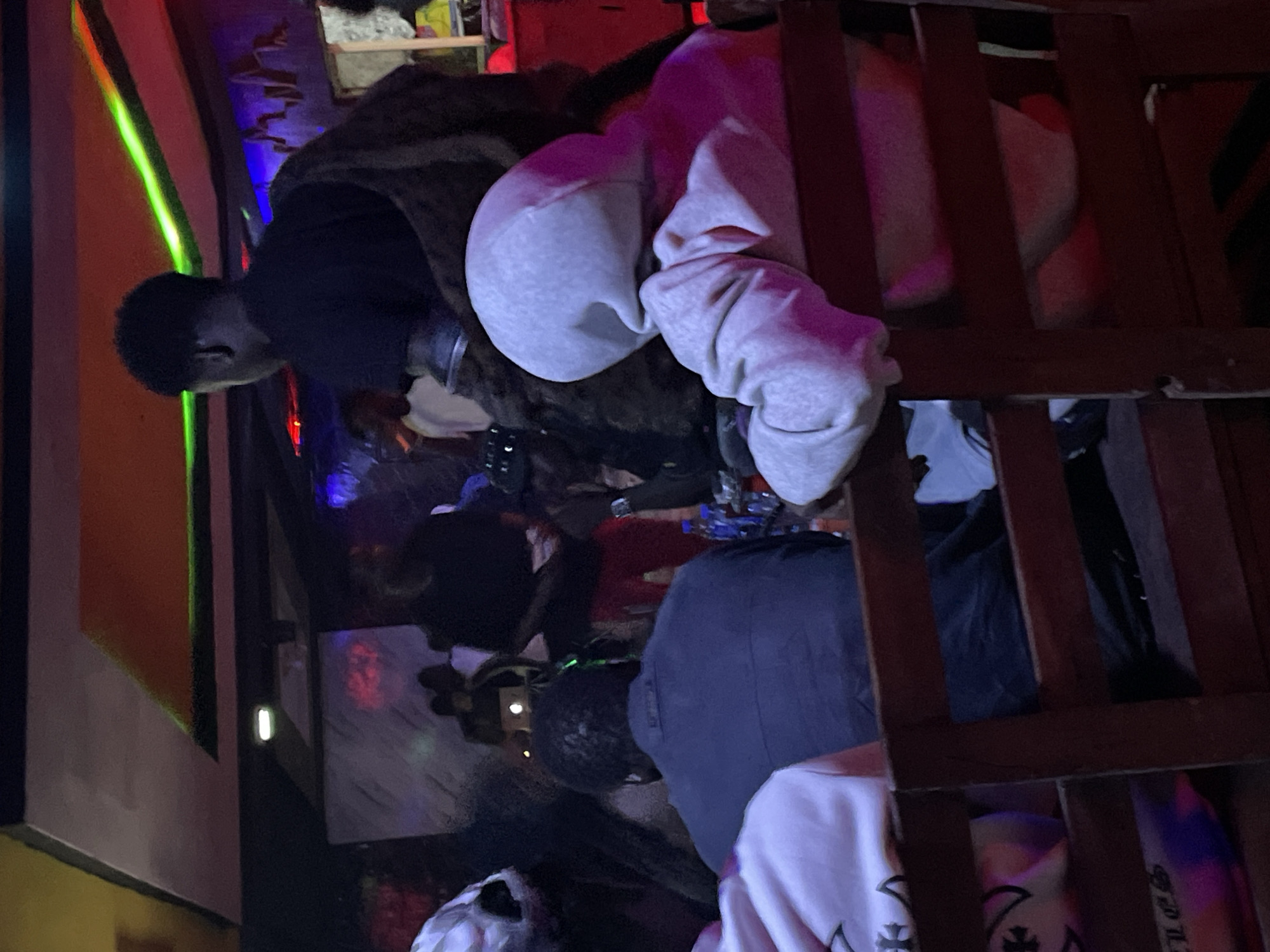 The image depicts an indoor scene, possibly a bar or nightclub, with dim, colorful lighting. Several individuals are visible, seated and standing, some facing away from the camera. In the foreground, two people are seated, one wearing a light-colored hooded sweatshirt and another wearing a dark jacket. Behind them, other individuals and elements of the environment are discernible, including a DJ booth or stage area with speakers and lighting. The overall atmosphere suggests a social gathering. The location is identified as Durumi, Nigeria.