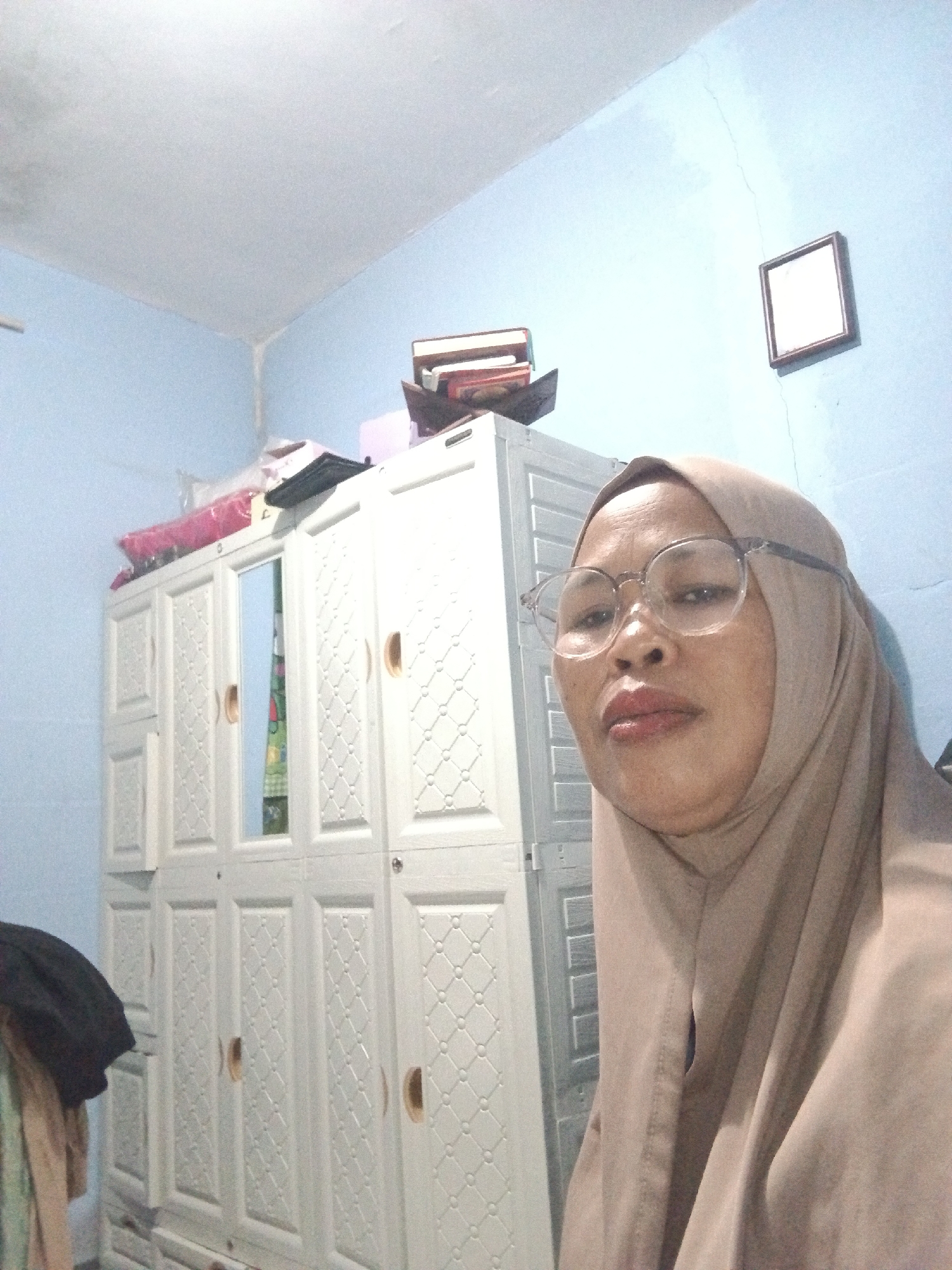 A medium shot captures a woman wearing a light brown hijab and round glasses, facing the camera directly. She has tan skin and full lips, and appears to be indoors, possibly in a bedroom or living area in Warungkiara, Indonesia. Behind her is a tall, white, multi-door wardrobe with textured detailing on each door. On top of the wardrobe, there are several stacked books and some pink and white plastic bags. The wall behind the wardrobe is painted a light blue, and a small, square framed picture hangs on it. The ceiling is white and appears to have a textured finish. To the left of the wardrobe, some dark clothing is visible, draped over what appears to be the edge of another piece of furniture. The overall lighting suggests an indoor environment with artificial light sources.