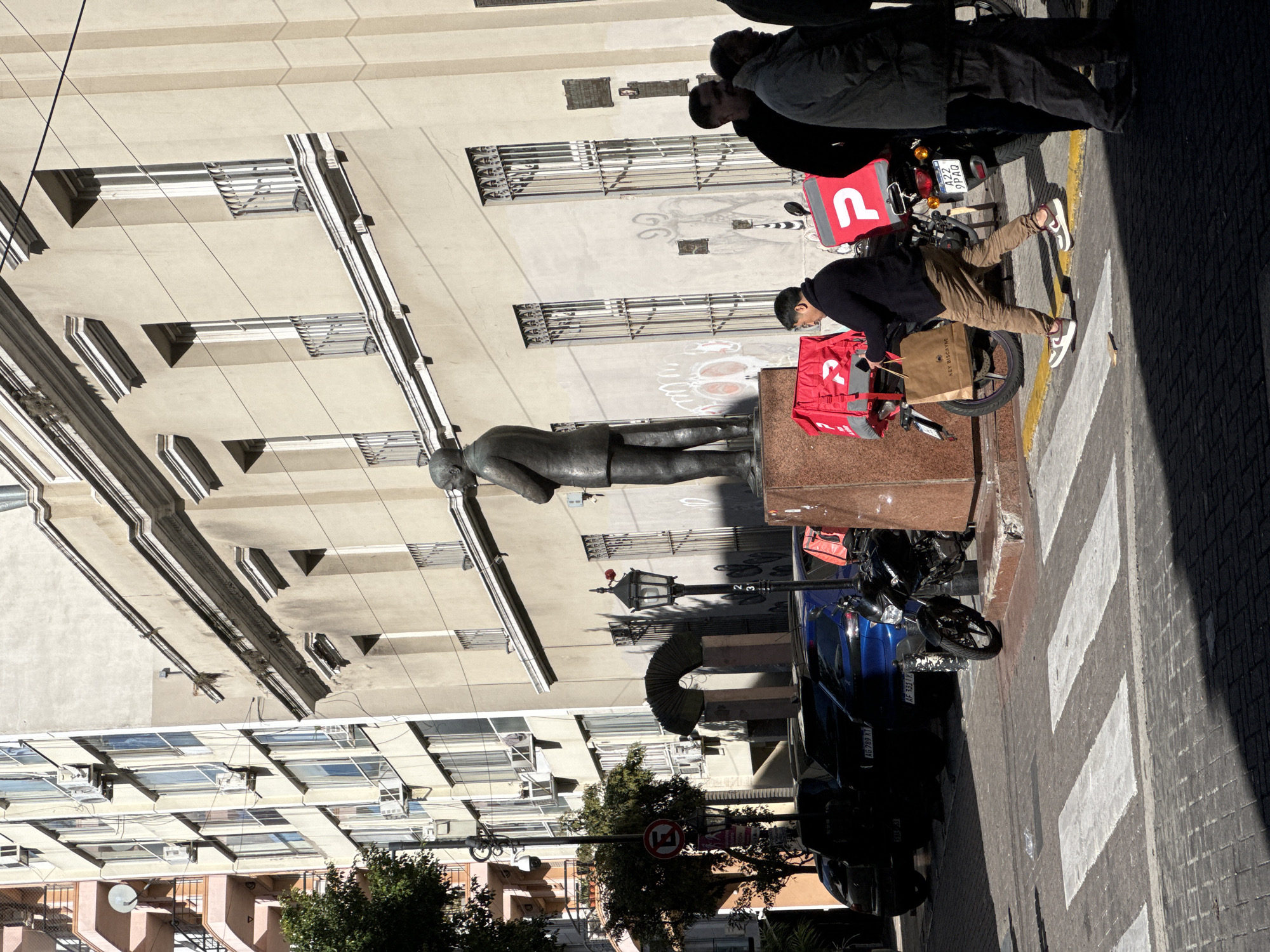 The image captures an outdoor scene in Buenos Aires, Argentina. In the foreground, a bronze statue of a man stands on a stone pedestal. Next to the statue, there are two delivery motorbikes with bright red delivery boxes, one belonging to "PedidosYa." Two delivery men are present: one is actively adjusting something on his bike, and the other is leaning near his bike. The street includes a blue car parked along the curb, as well as a motorcycle. Pedestrian crosswalk markings are visible on the pavement. The backdrop is a building with multiple windows and architectural details. A traffic sign with a "no parking" symbol is also visible near a green tree.