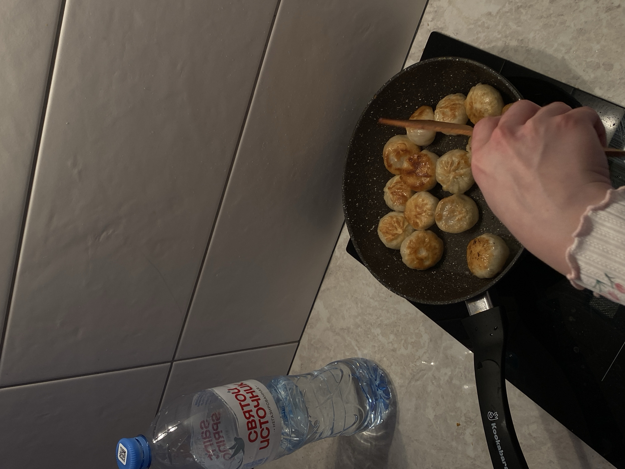 The image shows a pan with dumplings being cooked on a stovetop. A hand is using chopsticks to move the dumplings. A bottle of "Святой Источник" water is on the counter near the stovetop. The location is Зубово, Russia.