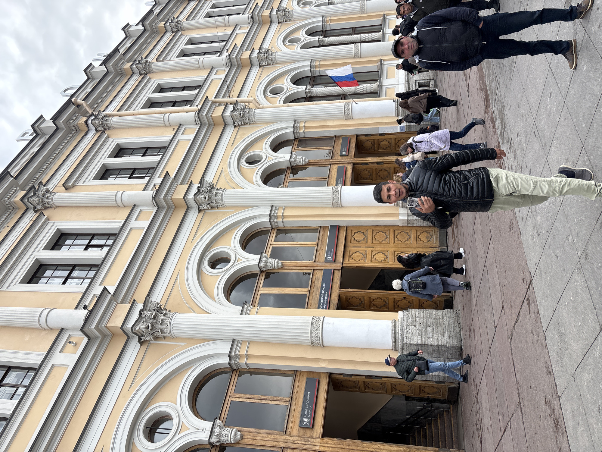 The image depicts the facade of the Moskovsky railway station in Saint Petersburg, Russia. The architectural style includes pale yellow walls, white arched windows, and white columns. A Russian flag hangs between the arched windows near the entrance. People are walking on the sidewalk in front of the station. Two men are facing the camera, one holding a mobile phone. A sign in Russian, translated to "No Entry", is visible on one of the entrance doors. The sky is overcast.