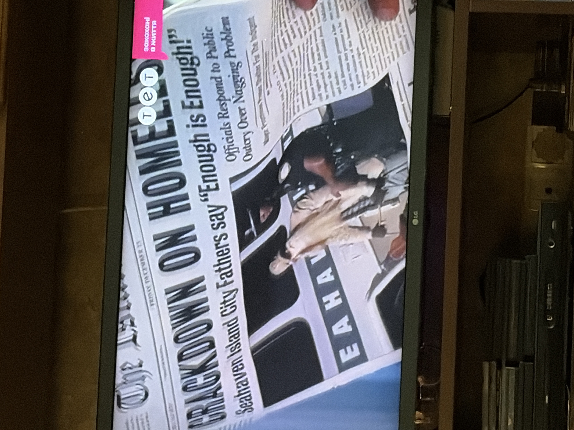 The image shows a television screen displaying a news article from a newspaper. The headline reads "CRACKDOWN ON HOMELESS". There is a smaller headline that says "Seahaven Island City Fathers say 'Enough is Enough!'". The article is printed in black ink on what appears to be a white or off-white newspaper. Below the headlines, there is an image of a police officer in uniform, appearing to interact with a person. The person is partially obscured and appears to be wearing civilian clothing. The background of the newspaper article features a graphic of a bus or train with the letters "E A H A V" visible. In the top right corner of the screen, there are three circular icons with the letters "т е т" inside them, and a pink speech bubble with Ukrainian text "закохані в життя" which translates to "in love with life". The television is placed on a shelf, and a stack of DVDs or Blu-rays is visible below it. The overall setting appears to be a domestic environment, likely a living room. No specific location in Osolynka, Ukraine is visible or suggested by the image content.