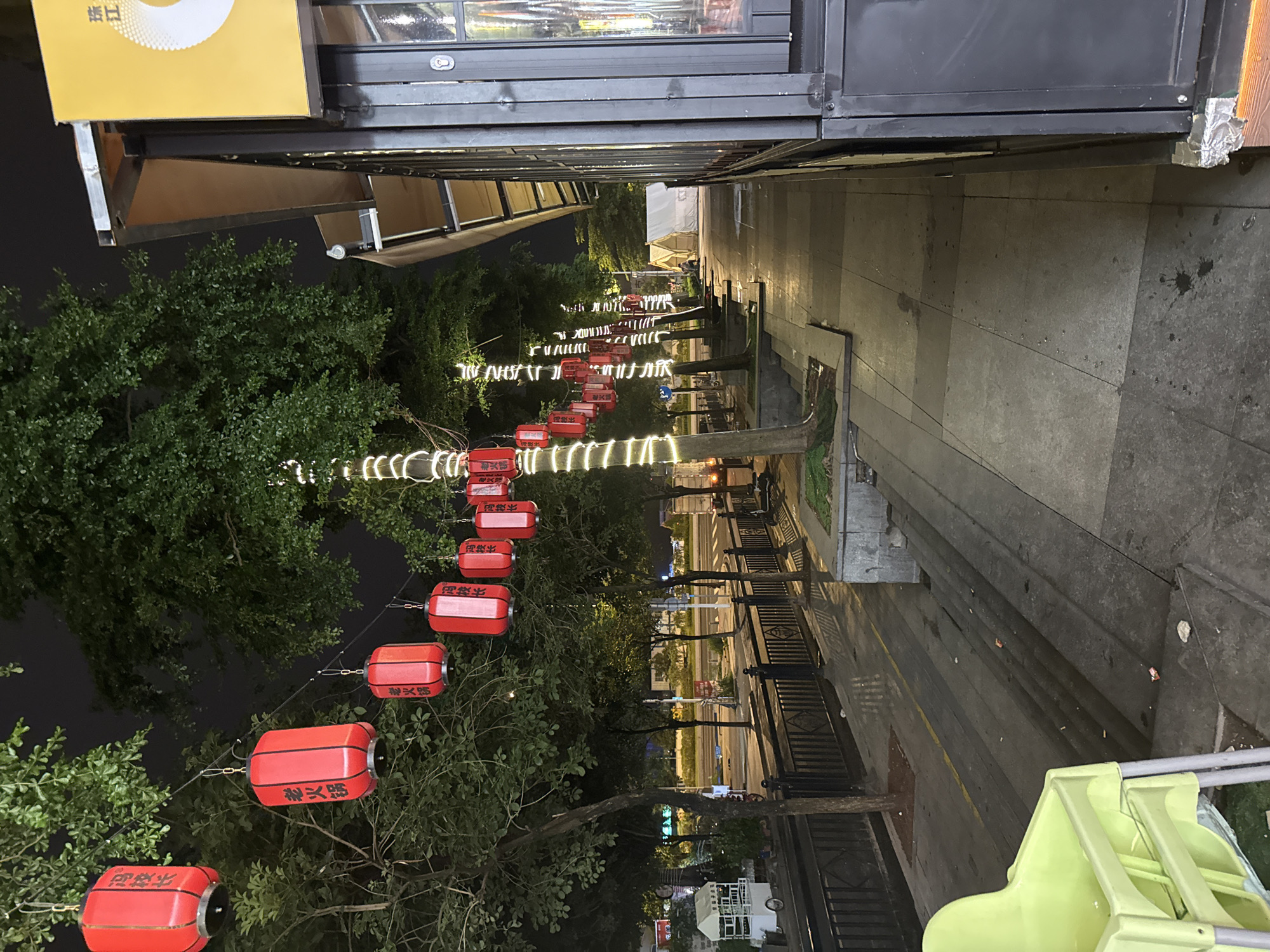 This image captures a nighttime street scene in Haizhu District, China, likely a commercial or entertainment area. Red lanterns, characteristic of Chinese celebrations, are strung across trees and poles lining the sidewalk. Some of these lanterns appear to be decorated with text in Chinese characters, suggesting they are related to local businesses or events. The trees themselves are adorned with strings of white fairy lights, adding a festive ambiance to the dark night.
To the right of the frame, a building with a dark metal facade and a yellow awning is visible. This building seems to be a shop or restaurant, with a glass window displaying interior lighting. The building's exterior suggests a modern, possibly commercial establishment.
The sidewalk in the foreground is paved with large, grey tiles, some of which are wet, indicating recent rain or street cleaning. A bright green child's high chair is prominently placed on the left side of the sidewalk, near some steps. Further down the sidewalk, illuminated tents are set up, suggesting temporary accommodations or stalls. A street runs alongside the sidewalk, with streetlights visible in the distance, casting reflections on the wet road. The overall impression is a lively, yet quiet, urban setting illuminated by artificial lights and decorated with traditional and modern elements.