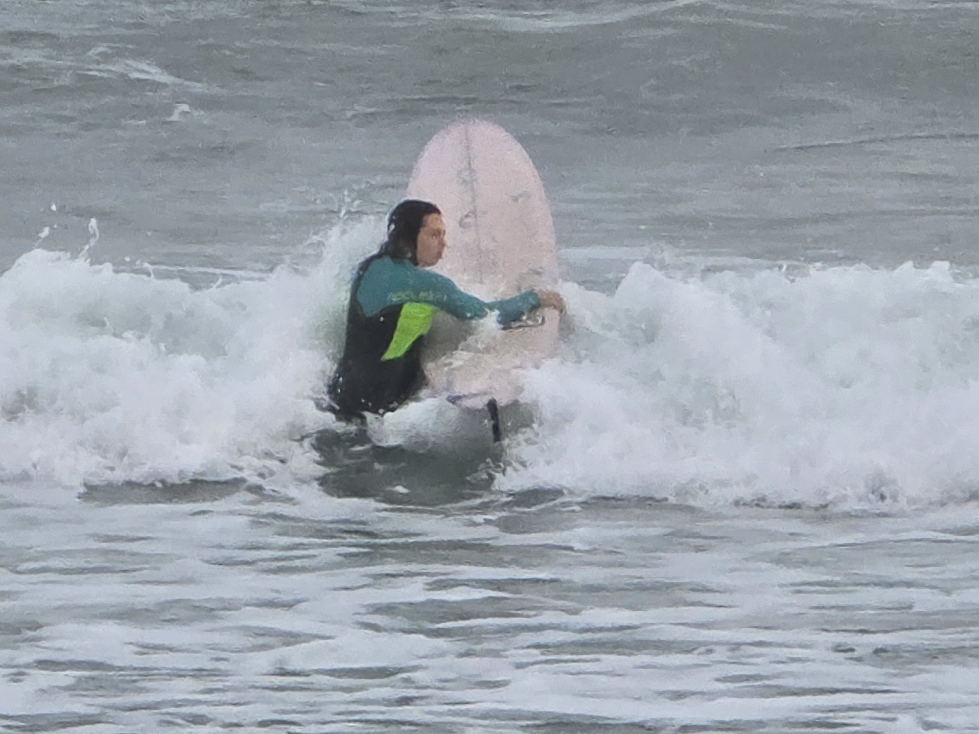 A person in a wetsuit stands in the ocean with a surfboard, braced against a small wave. The person has dark hair and is looking to the left of the frame. The wetsuit is black with a teal upper sleeve and a bright green side panel. The surfboard is light pink and positioned vertically in the water. The ocean water is grey, with visible waves and foam. The scene suggests the sport of surfing, potentially taking place at a beach in Tel-Aviv, Israel.
