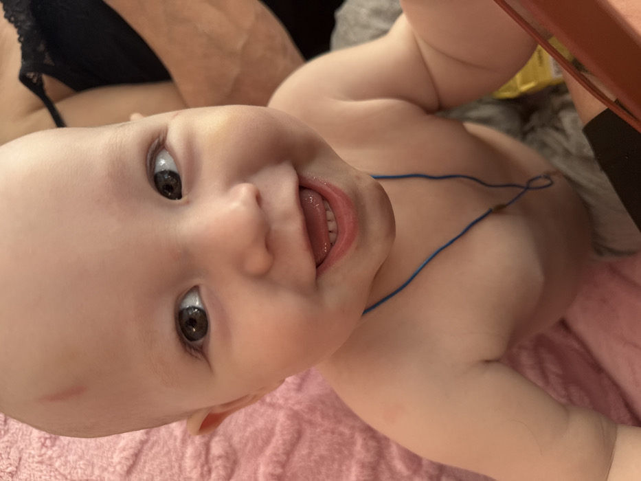 The image displays a close-up, eye-level portrait of an infant or toddler with fair skin, captured indoors in the city of Кипчак, Russia. The child, visible from the chest upwards, is bare-chested and has dark eyes, with a wide, open-mouthed smile revealing two lower and two upper front teeth. A faint vertical reddish mark is present on the upper left forehead. A thin blue string necklace with a small dark bead is worn around the child's neck. The child is looking slightly upward and to the viewer's left. The background on the left is a textured pink fabric. On the right, a partially visible adult in a black lace garment is present, with an arm also discernible. In the bottom right corner, a reddish-brown object, possibly a mobile device, and faint glimpses of grey fabric and a yellow object are visible.