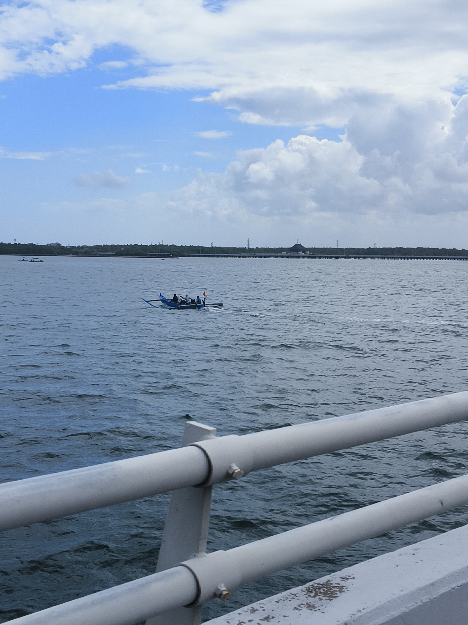 The image depicts a body of water, likely the sea, under a partly cloudy sky. In the foreground, a white metal railing, part of a bridge or pier, runs diagonally across the frame. The water is choppy, indicating some wind.
Further out on the water, a small blue boat with an outrigger is visible. Several people are seated in the boat. One person appears to be steering, while others are observing or resting. A small orange flag is attached to the stern of the boat. In the distance, a treeline marks the shore, with some structures and power lines visible beyond it. A long, elevated structure, possibly a bridge or causeway, stretches across the horizon.
The scene suggests a typical day in a coastal area. Given the description of Kuta Selatan, Indonesia, this scene could be from a location like Jimbaran Bay, known for its beaches, seafood restaurants, and boat activity. The boat's design, with outriggers, is characteristic of traditional Indonesian fishing boats. The presence of the railing in the foreground implies the vantage point is from a man-made structure overlooking the water. The lighting suggests it is daytime, possibly late morning or early afternoon, with the sun not being directly overhead.