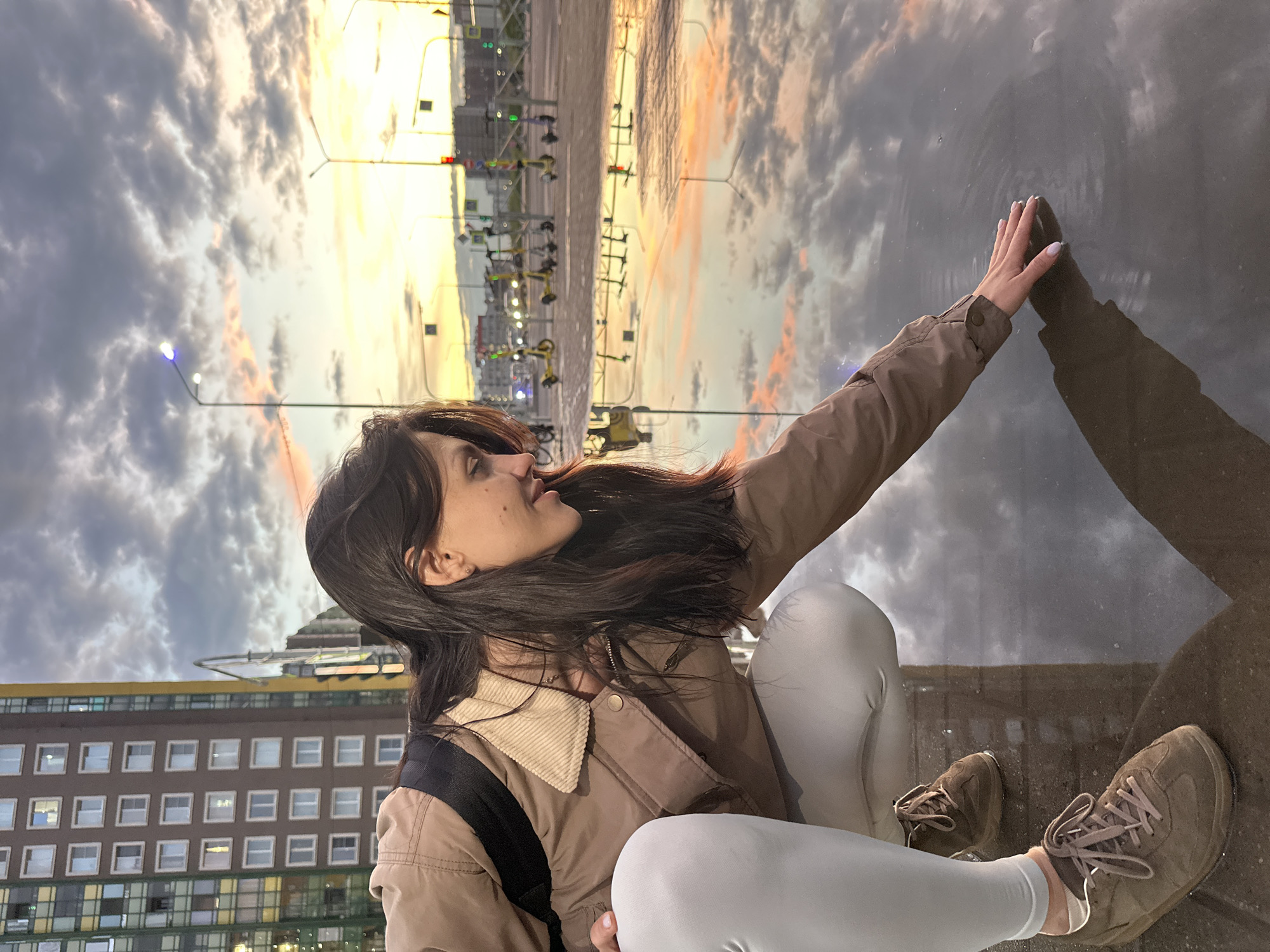 In the city of Unknown, Unknown, a young woman with long, dark hair crouches down to touch a puddle of water. She is wearing a brown jacket over a white shirt and light-colored leggings, with brown athletic shoes. Her right hand is extended, fingers lightly touching the surface of the water, creating ripples. The puddle reflects the cloudy sky, which is illuminated by the warm hues of a sunset. In the background, a tall, modern building with numerous windows stands to the left. Further back, streetlights and traffic signals are visible, along with several parked electric scooters. The overall scene suggests an urban environment during twilight.