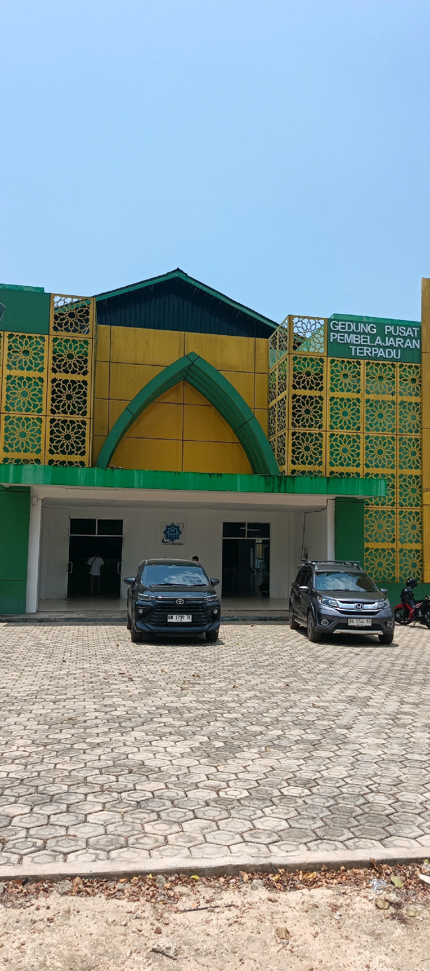 This is a full-shot, eye-level, vertical photograph of the exterior of a building on a sunny day. The building has a distinct architectural style with a yellow facade and green accents. Above the entrance, there is a prominent sign that reads "GEDUNG PUSAT PEMBELAJARAN TERPADU." The building's facade is adorned with decorative green panels featuring intricate geometric patterns.
In front of the building, there is a paved courtyard area made of hexagonal concrete blocks. Two cars are parked in front of the entrance. The car on the left is a black SUV, and the car on the right is a dark grey SUV. There are also two motorcycles parked to the right of the grey SUV, one red and one black.
A person in a white shirt is visible through the left entrance of the building. Another person, partially obscured, is standing near the right entrance.
The sky is clear and blue, suggesting good weather conditions. The ground in the foreground is a dirt path with some dried leaves and debris scattered on it. The overall scene appears to be a public building, possibly a school or a community center, located in Desa Namang, Indonesia.