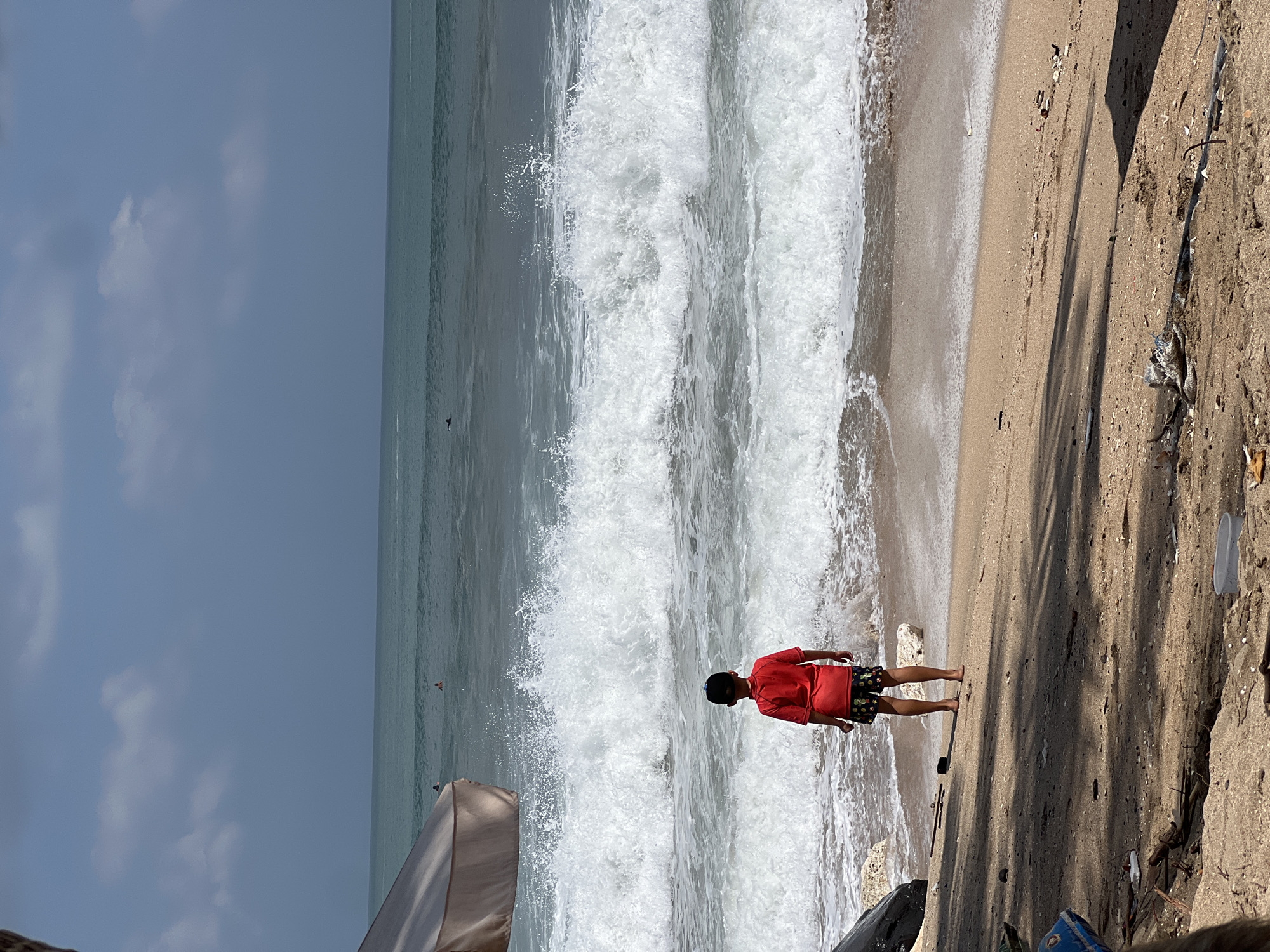 A lone child, wearing a red shirt and patterned shorts, stands on the sandy shore of Kuta Beach, Bali, Indonesia, facing a large wave about to crash. The child's back is to the viewer, and they appear to be contemplating the powerful ocean. The beach is littered with debris, including driftwood, plastic bottles, and other refuse, indicating a potential environmental issue. In the background, the vast ocean stretches to the horizon under a partly cloudy sky. Several surfers are visible in the distance, enjoying the waves. A large umbrella shades a portion of the beach in the foreground, casting shadows on the sand. The overall scene captures a moment of quiet observation amidst the dynamic energy of the sea.