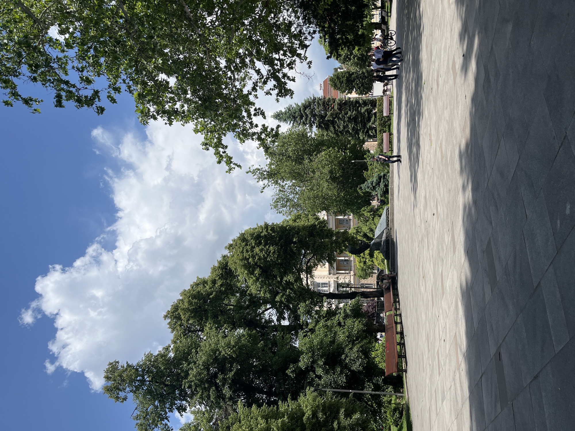 The image shows a section of a park in Sofia, Bulgaria. The foreground consists of a paved area with trees lining the sides, casting shadows on the paving. Benches are situated along the side of the paved area, near the trees. A person is walking along the paved area. In the background, buildings are visible amongst the trees. The sky above has blue patches and white clouds.