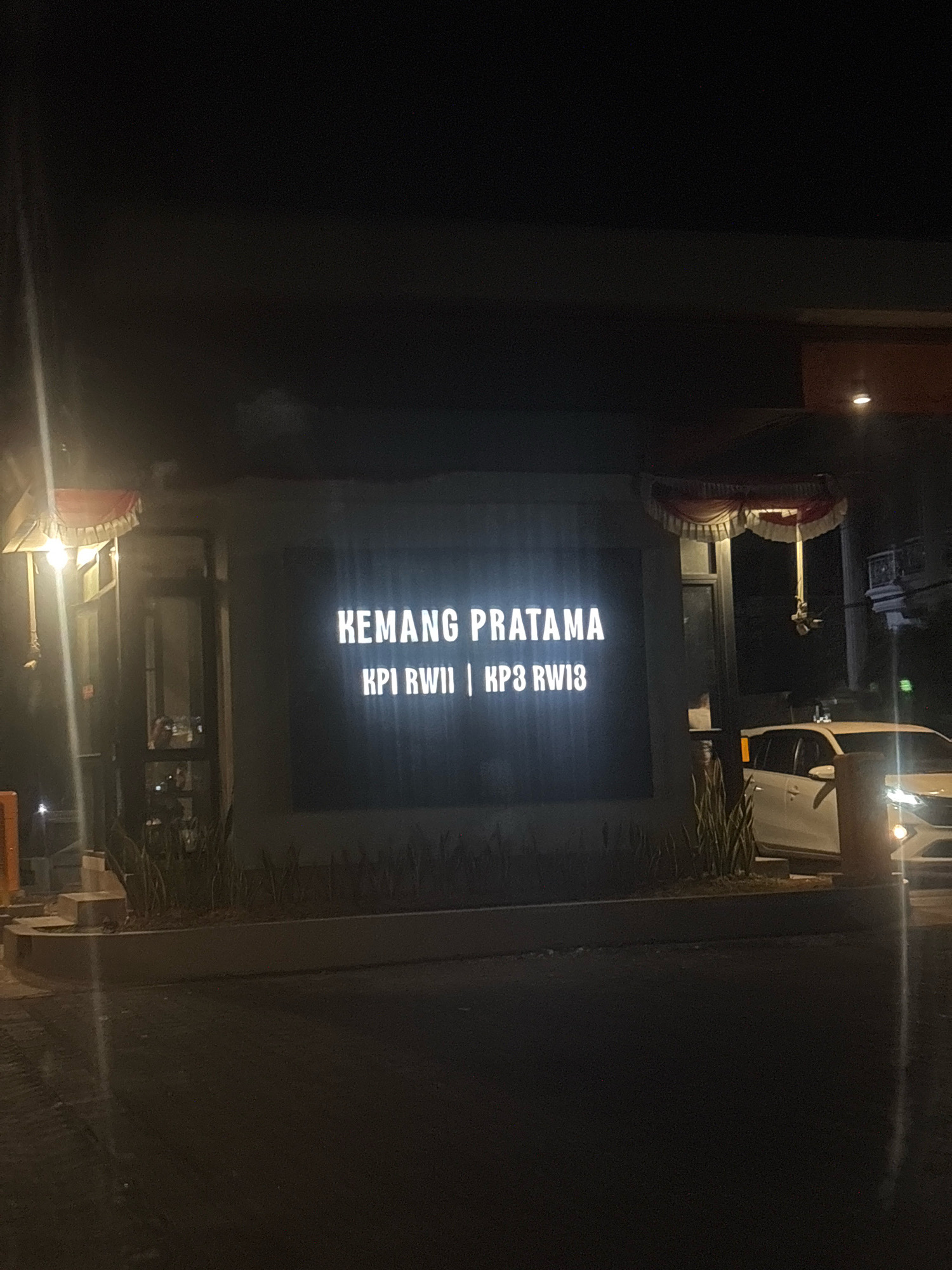 A nighttime exterior shot shows a lit sign that reads "KEMANG PRATAMA KPI RWII | KP3 RW13". The sign is mounted on a building facade with decorative awnings above it. To the right of the sign, a white SUV is partially visible. The surrounding area appears to be a street or driveway, with dark pavement and some landscaping. The lighting suggests it is an urban or suburban environment, likely an entrance to a residential complex or business park. The image is taken from a low angle, looking up towards the sign. The city of Bekasi, Indonesia is a possible location for this scene, given the common use of "Kemang Pratama" as a name for residential areas in Indonesia.