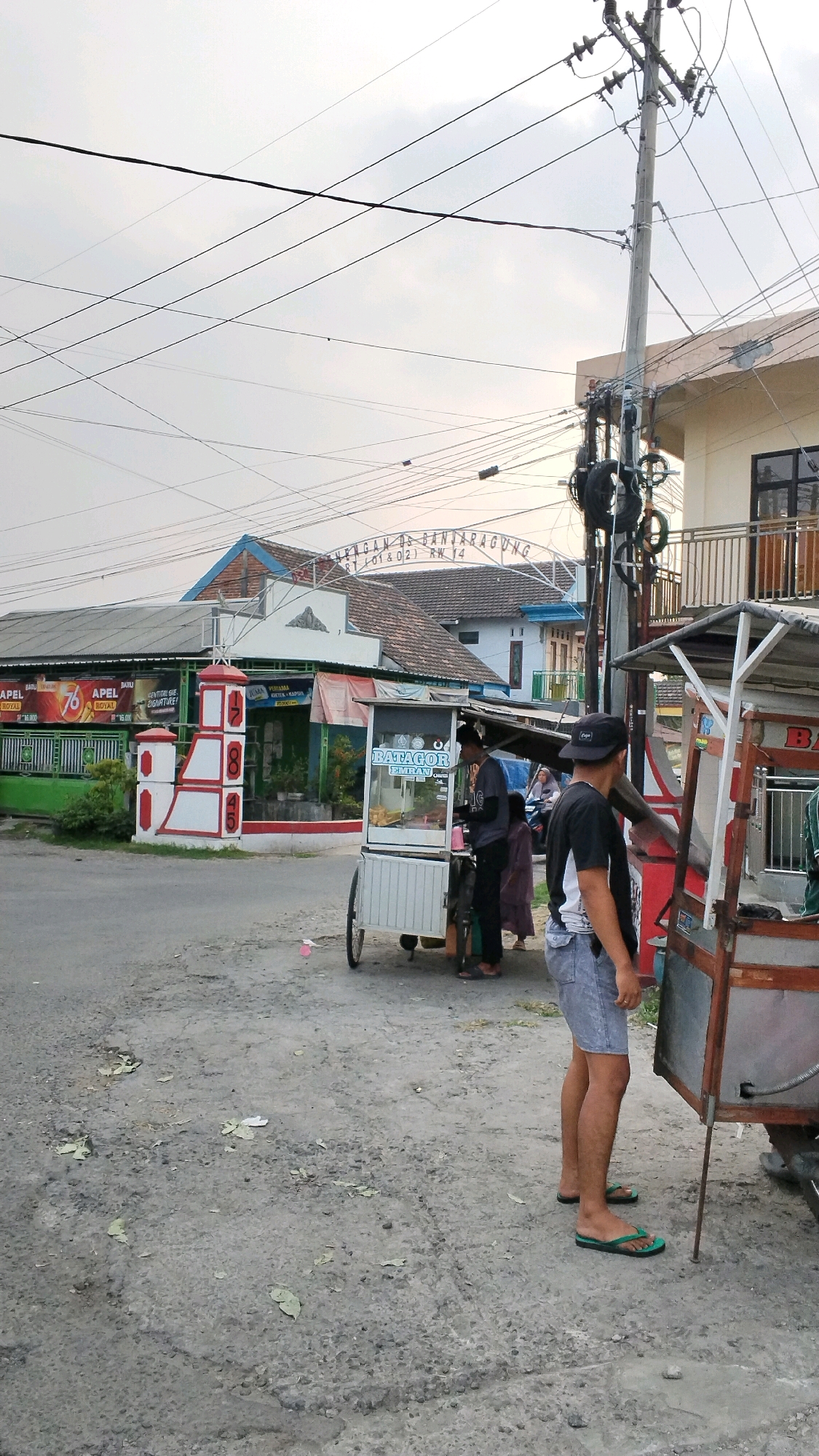 The image captures a street scene in Banjaragung, Indonesia, likely during daytime given the lighting. In the foreground, a man wearing a black cap, a black and white t-shirt, and light denim shorts stands facing a food cart. He is wearing green flip-flops. The food cart has a white base with a covered top and appears to be selling some kind of food item, with "BATAGOR" and "PIMRAN" visible on signage. To the left of this cart, another food cart is partially visible, with a man in a gray t-shirt and black pants attending to it. A young child in a purple garment stands near the second cart.
Behind the food carts, a street corner is visible. A building with a white facade and a red and white decorative pillar is on the left, with green fencing and advertising banners displaying "76 ROYAL" and "BARU." An arched sign overhead reads "SELAMAT DATANG DI BANJARAGUNG" and includes text about roads and an RN number. Further back, more buildings with tiled roofs are visible, and numerous electrical wires crisscross the sky, supported by a utility pole. The ground is a rough asphalt or concrete surface with scattered leaves. The overall impression is a typical roadside commercial area in a town or city.