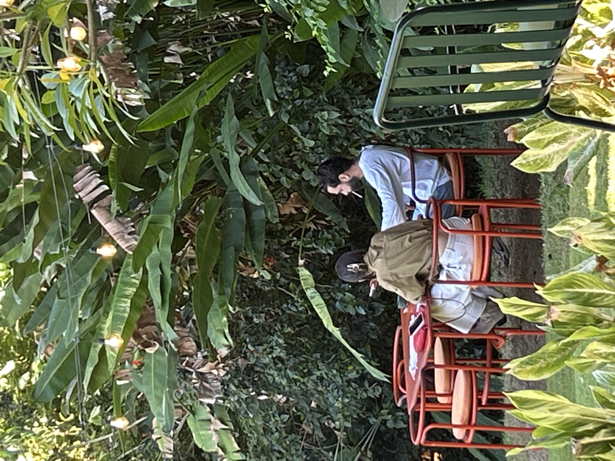 In Mengwi, Indonesia, two individuals are seated at a red table outdoors, surrounded by lush greenery and hanging string lights. A man wearing a white long-sleeved shirt is smoking a cigarette and facing left. A person with dark hair, wearing a tan jacket and white shorts, is seated opposite him, facing away from the camera and holding a phone. A red phone and a red wallet are on the table. The setting appears to be a relaxed, outdoor dining or seating area.