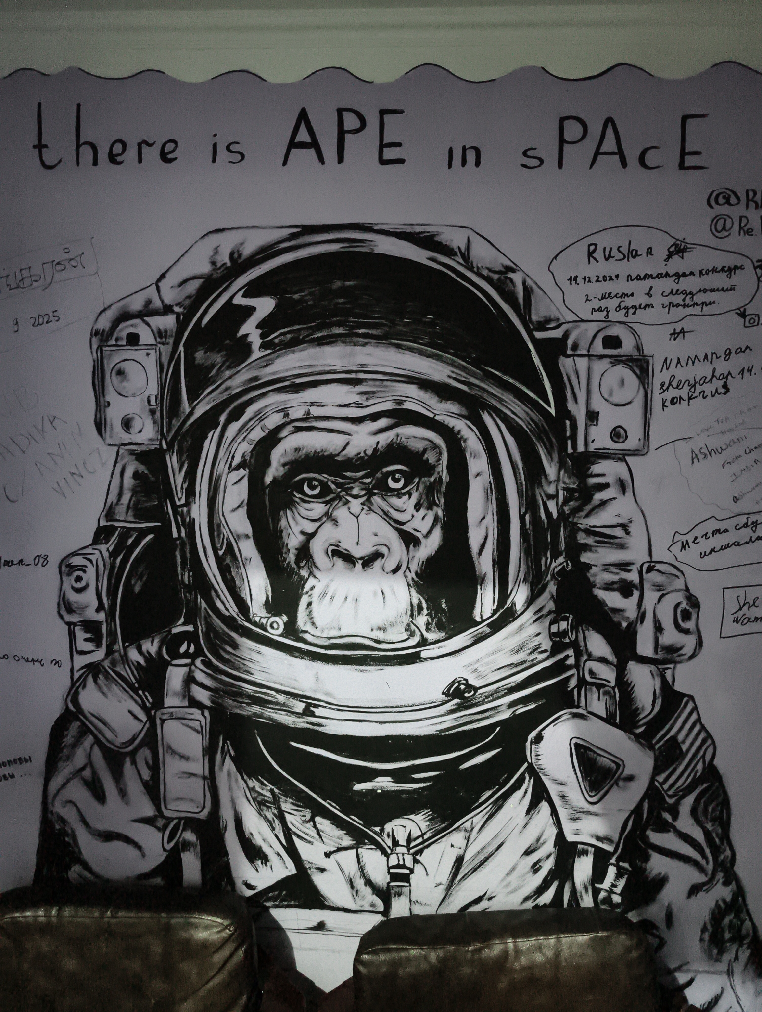 The image shows a black and white drawing of an ape wearing a space suit. Above the drawing, the phrase "there is APE in sPACe" is written in black marker. Surrounding the drawing and text are various signatures and notes written in different languages. Based on the text "NAMANgan sherjahap 14," the drawing is likely located in Namangan, Uzbekistan, as it refers to a sherjahap (wedding feast). However, this is NOT Tashkent, Uzbekistan, as specified in the instructions. The drawing is positioned behind a dark brown couch or seating area. The lighting is ambient, casting shadows on the lower part of the image. The style of the drawing is illustrative, with thick lines and shading used to create depth and detail. The background appears to be a wall, which is likely light-colored. The overall tone of the image is informal and artistic.