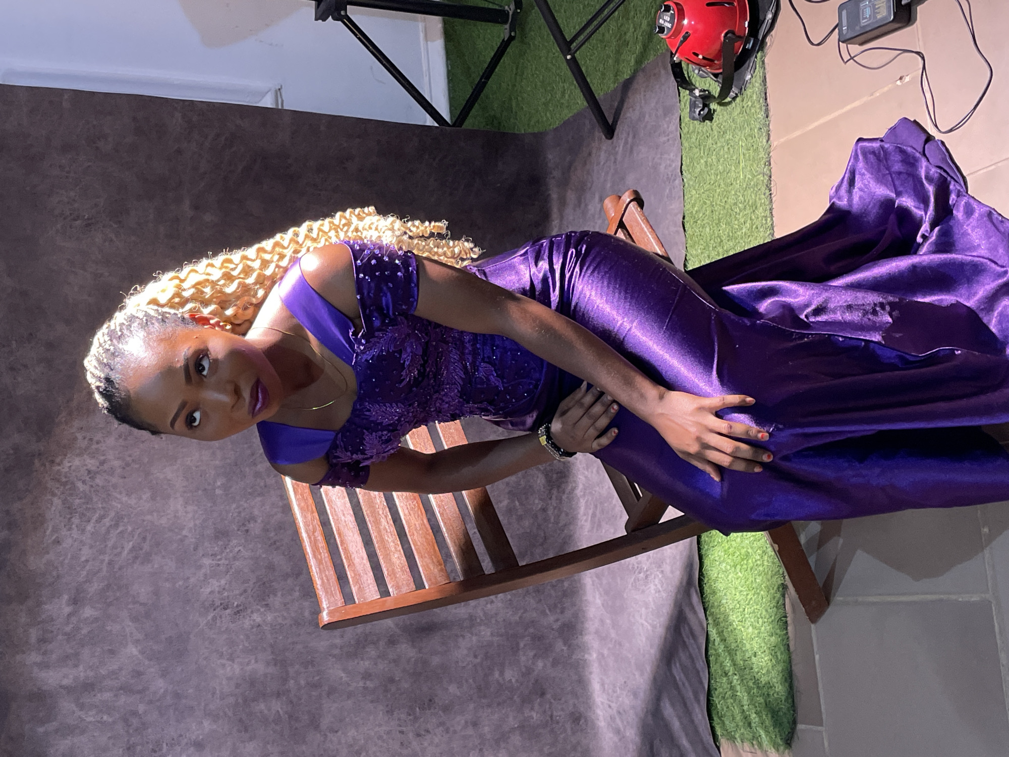 An indoor portrait captures a woman elegantly posing in what appears to be a photography studio in Barakin Akawo, Nigeria. The woman, of African descent, is seated sideways on a light brown wooden rocking chair, looking directly at the camera. She wears a striking purple gown, featuring an embellished, possibly sequined or embroidered, off-shoulder bodice and a flowing, shimmering satin-like skirt. Her long, voluminous blonde curly hair or braids are styled to cascade over her shoulder. She has a subtle necklace and a dark beaded bracelet on her left wrist, and her fingernails are painted red. Her expression is calm and composed, with noticeable eye makeup and purple lipstick.
The setting includes a mottled dark brown or grey backdrop behind her, providing a soft contrast to her vibrant dress. The floor transitions from a smooth, dark grey tile to a section of bright green artificial grass. Studio equipment is visible around her: a black light stand is partially seen at the top left, and a red studio light fixture with the text "LED 80W VAC" is positioned on the right side of the frame, along with an associated power adapter or controller with visible wires and some unreadable text. The lighting suggests a controlled studio environment, highlighting the woman and her dress.
