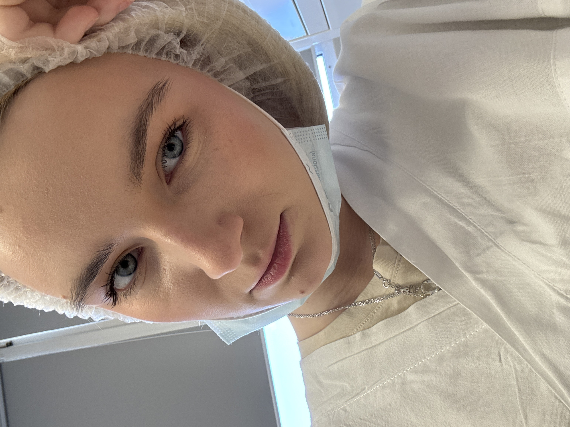 A person is looking down at the camera, wearing a white bouffant cap and a surgical mask pulled below their nose. They have fair skin, blue eyes, and light eyebrows and eyelashes. A silver necklace is visible around their neck. They are wearing a white shirt. The background appears to be a light gray or white surface with some indistinct light fixtures. The location cannot be identified as Saint Petersburg, Russia based on the given image.