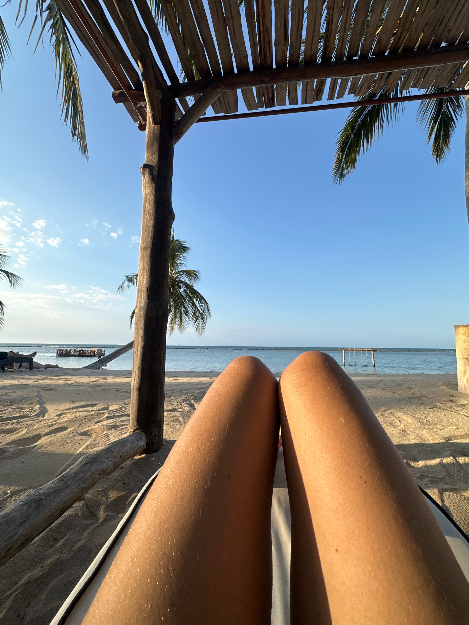 This is a first-person perspective shot of a person relaxing on a beach in Pasikudah, Sri Lanka. The foreground is dominated by the person's tanned legs, stretched out on what appears to be a lounge chair. Behind their legs, the sandy beach stretches towards the ocean. In the mid-ground, palm trees and a rustic wooden structure, possibly a canopy or shelter, are visible against a clear blue sky. The ocean is calm, and a wooden platform with a swing set is visible in the distance. The overall atmosphere is one of relaxation and vacation.