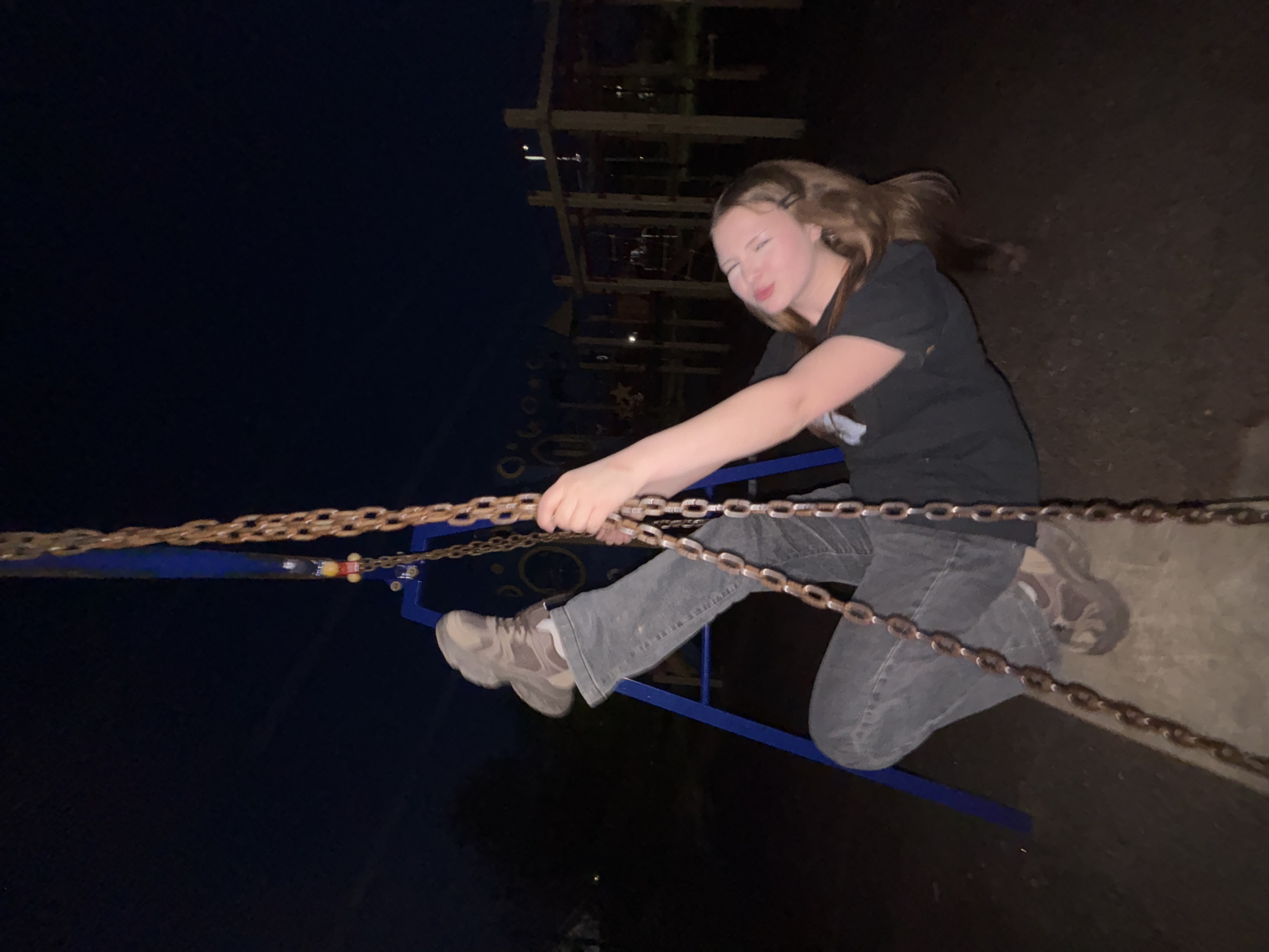 A person, likely a teenage girl, is suspended in the air, apparently swinging. She is gripping a chain with both hands and has a playful expression on her face with her eyes squinted. She is wearing a dark t-shirt, gray jeans, and light-colored shoes. She is positioned above what appears to be a blue-painted metal structure that is connected to the chain she is holding. In the background, there is a taller, more elaborate metal structure. The surrounding environment appears dark, suggesting it is nighttime. The location is Фоки, Russia, based on the metadata.