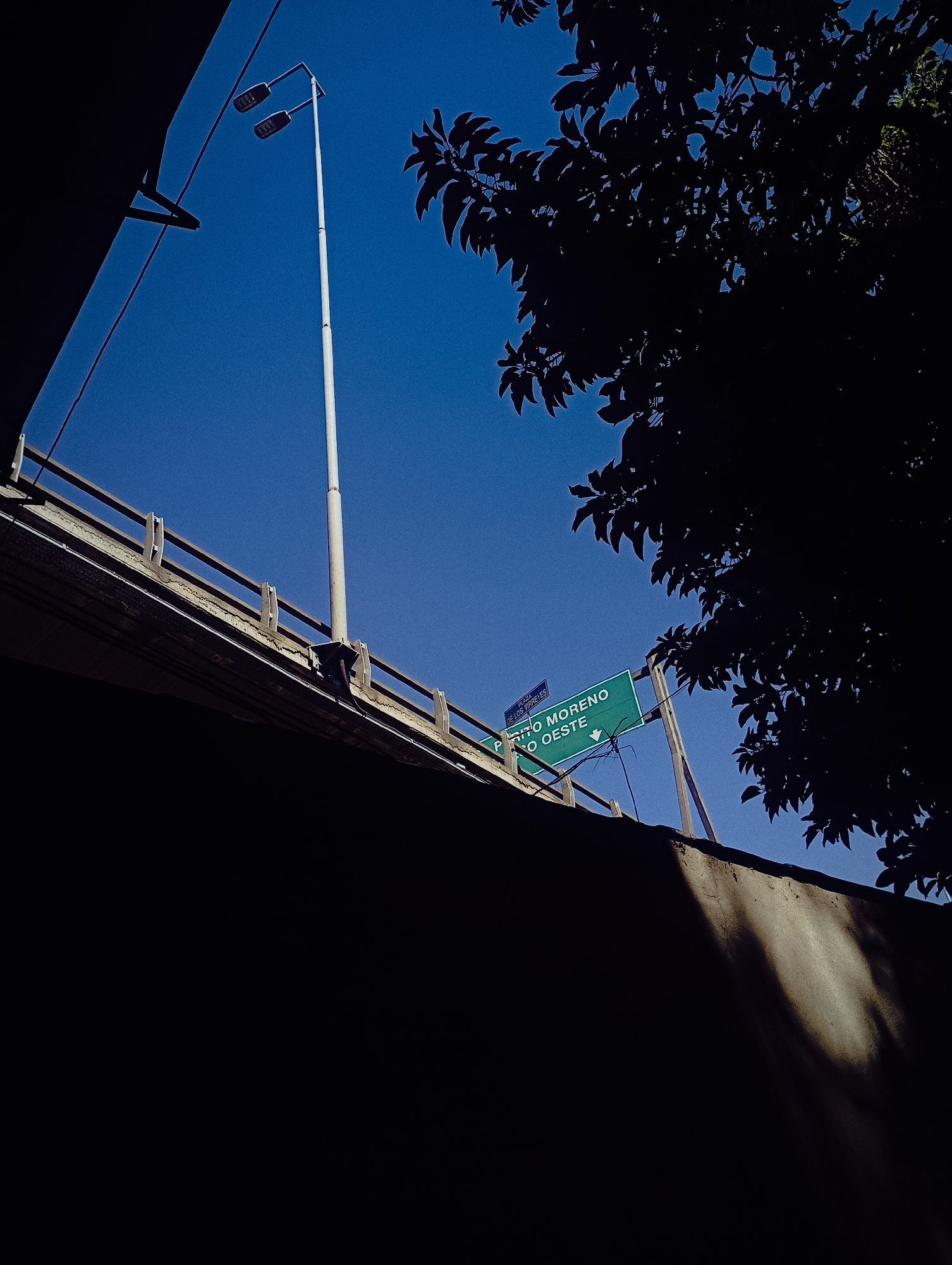 The image depicts an upward perspective of a bridge structure in Buenos Aires, Argentina. A highway sign indicates "Puerto Moreno," directing traffic towards "Rio Oeste" with a downward-pointing arrow. A tall streetlight stands nearby. The scene includes tree foliage that partially obscures the view. The foreground is dominated by a dark, shadowed structure. The sky is a clear, deep blue.
