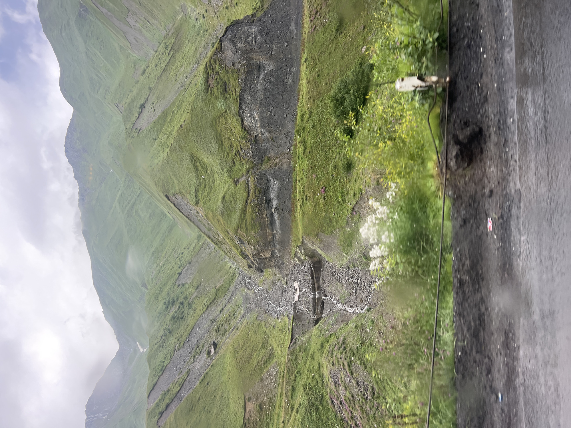 The image depicts a mountainous landscape in Almasiani, Georgia. The scene is dominated by lush green, undulating hills, some of which show evidence of rock slides or erosion, with exposed dark gray rock formations. A stream or small river carves a path down a rocky ravine in the center of the image, with water visible cascading over some ledges. Wildflowers in shades of pink and white dot the grassy slopes. In the foreground, a dark, rough surface, likely a road or embankment, is visible, with a dark wire running horizontally across it. A white post or marker is also present near the right edge of the foreground. The sky is overcast with clouds. There are no people or animals visible in the image.
