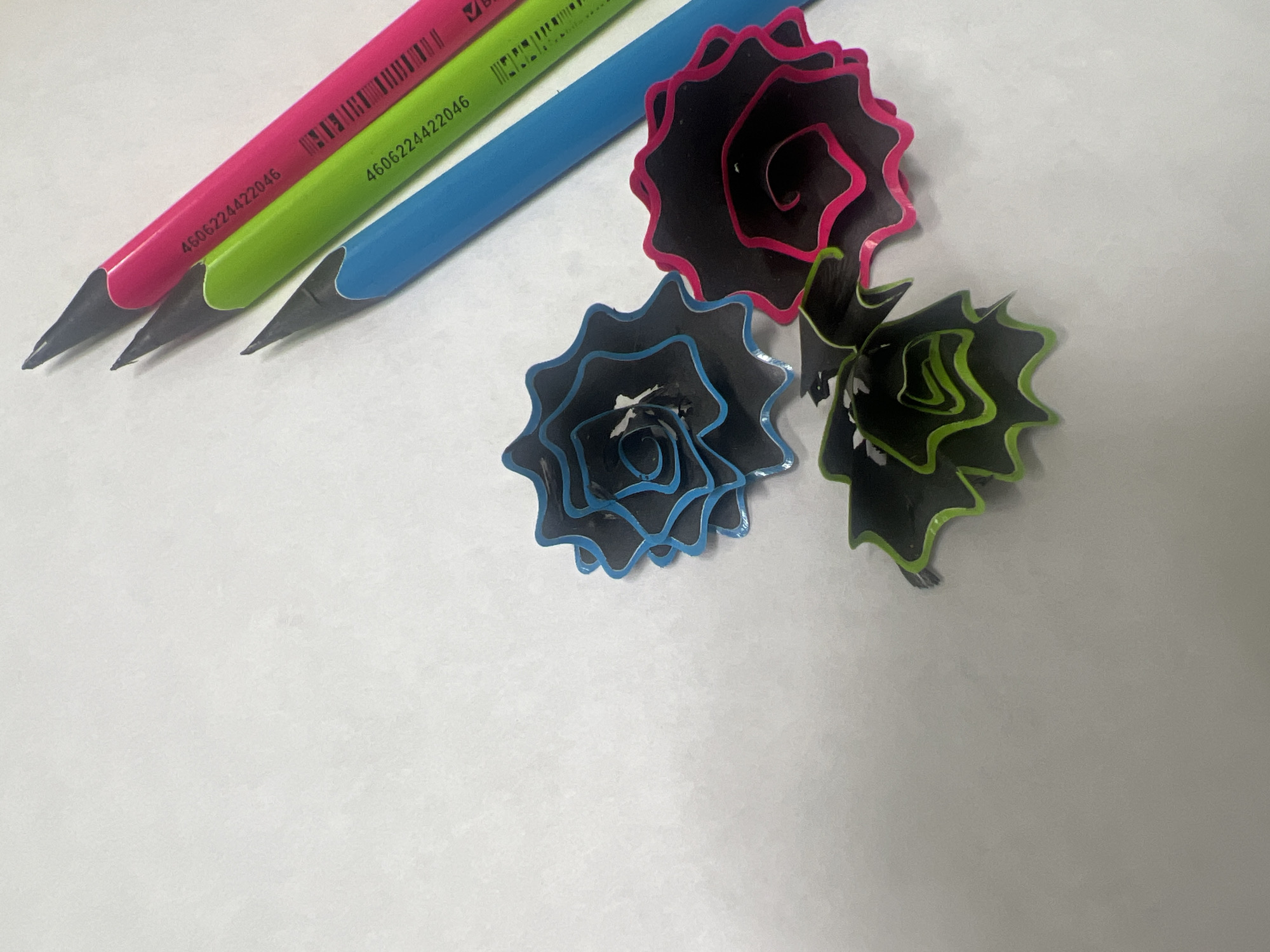 The image shows three sharpened pencils (pink, green, and blue) lying on a white surface, alongside three pencil shavings shaped like roses, each corresponding in color to one of the pencils. A numerical code is visible on the pink and green pencils. The location in Chaikovsky, Russia is not discernible from the image. There are no people present in the image.