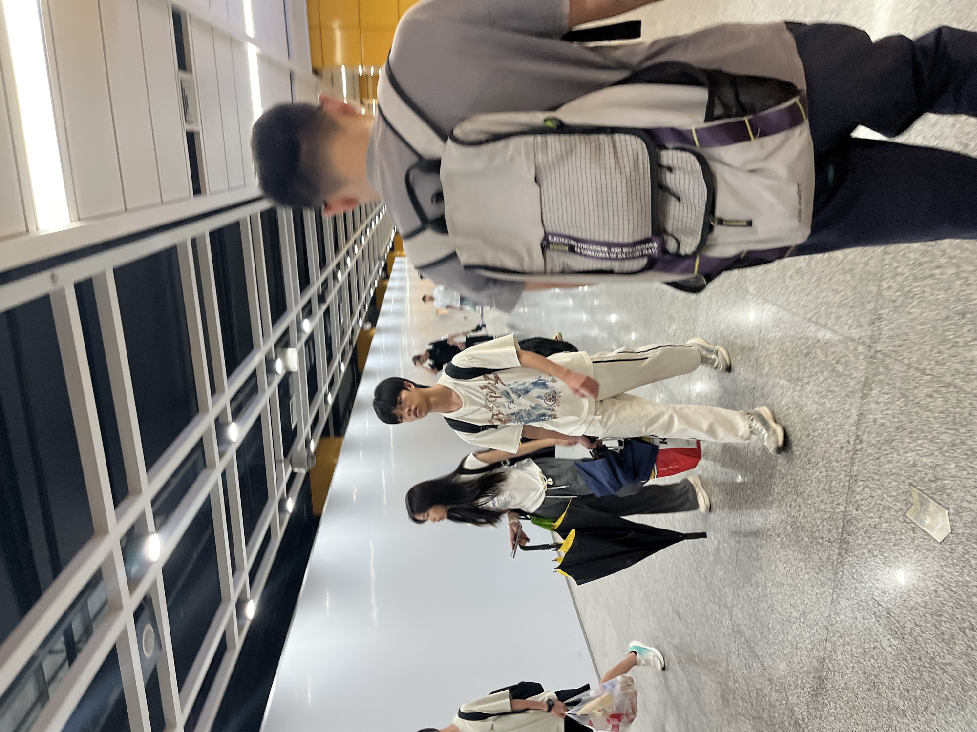 The image captures a dimly lit, indoor hallway with a polished concrete floor, likely a transportation hub or public building in Panyu District, China, given the context. Several individuals are walking away from the camera. In the foreground, a person wearing a grey t-shirt and dark pants walks away, carrying a light-colored backpack with purple accents. To their left, a young person in a white t-shirt and light-colored pants walks with a backpack. Further left, a young woman with long dark hair, also wearing a white top and grey pants, walks while holding a black umbrella and a bag. Another individual, partially visible on the far left, wears a light-colored t-shirt and shorts and carries a plastic bag. The ceiling features a grid of white panels and dark recesses, with embedded lighting. Yellow accents are visible in the background on the right side of the hallway. A small, metallic-looking object lies on the floor in the midground.
