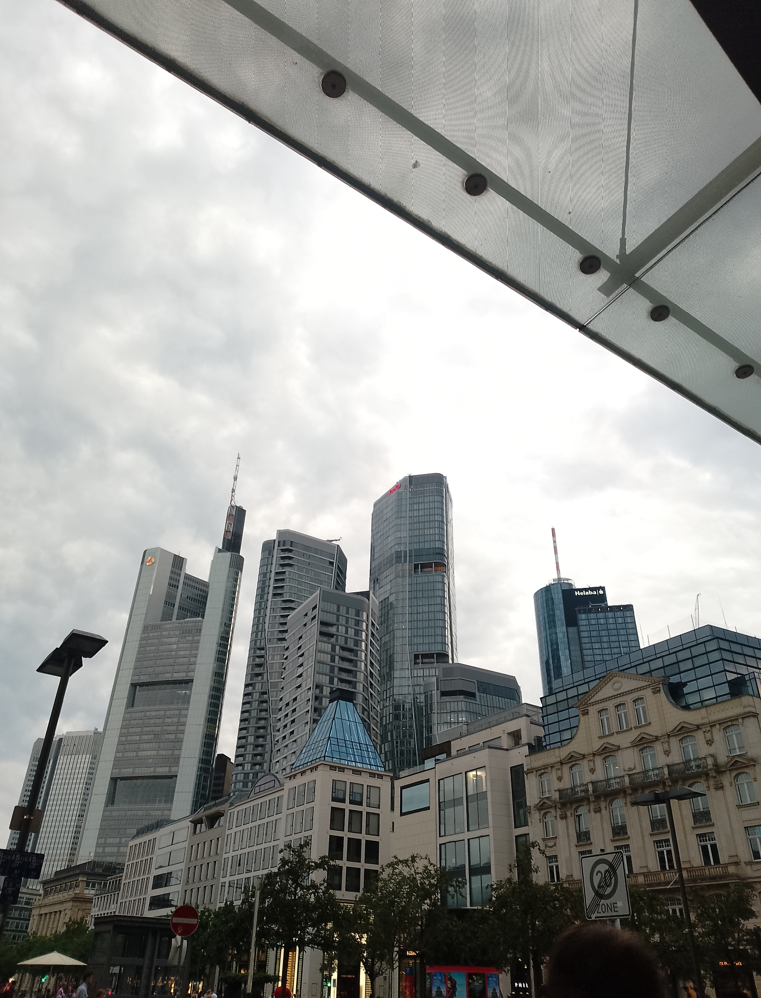 The image shows a low-angle view of a city skyline dominated by modern skyscrapers under a cloudy sky. In the foreground, a portion of a translucent, possibly metallic, overhead structure with visible rivets and supports occupies the top of the frame. Several skyscrapers of varying heights and architectural styles are visible. One prominent building features the logo of Commerzbank. Another skyscraper has "Helaba" written on it. In the middle ground, there are lower buildings with classical architectural elements, including a building with arched windows and ornate detailing. Trees line the street in front of these buildings. A street lamp is visible to the left. A traffic sign with a red circle and a horizontal bar, indicating a prohibition, and a "ZONE" sign are also present. People are visible in the lower part of the image, appearing to walk on a street or sidewalk. The overall impression is of a bustling urban environment with a mix of contemporary and older architecture.