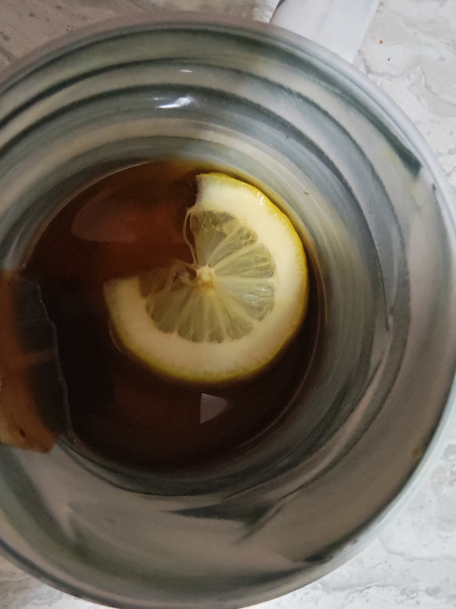 A close-up overhead view shows a slice of lemon floating in a dark liquid, likely tea or coffee, within a ceramic mug. The mug has a distinctive blue and grey swirled pattern on its exterior. The background is a light-colored, textured surface, possibly a marble countertop. There are no people visible in the image. The scene is indoors, likely in a kitchen or dining area. No specific location in Tolyatti, Russia, can be identified.