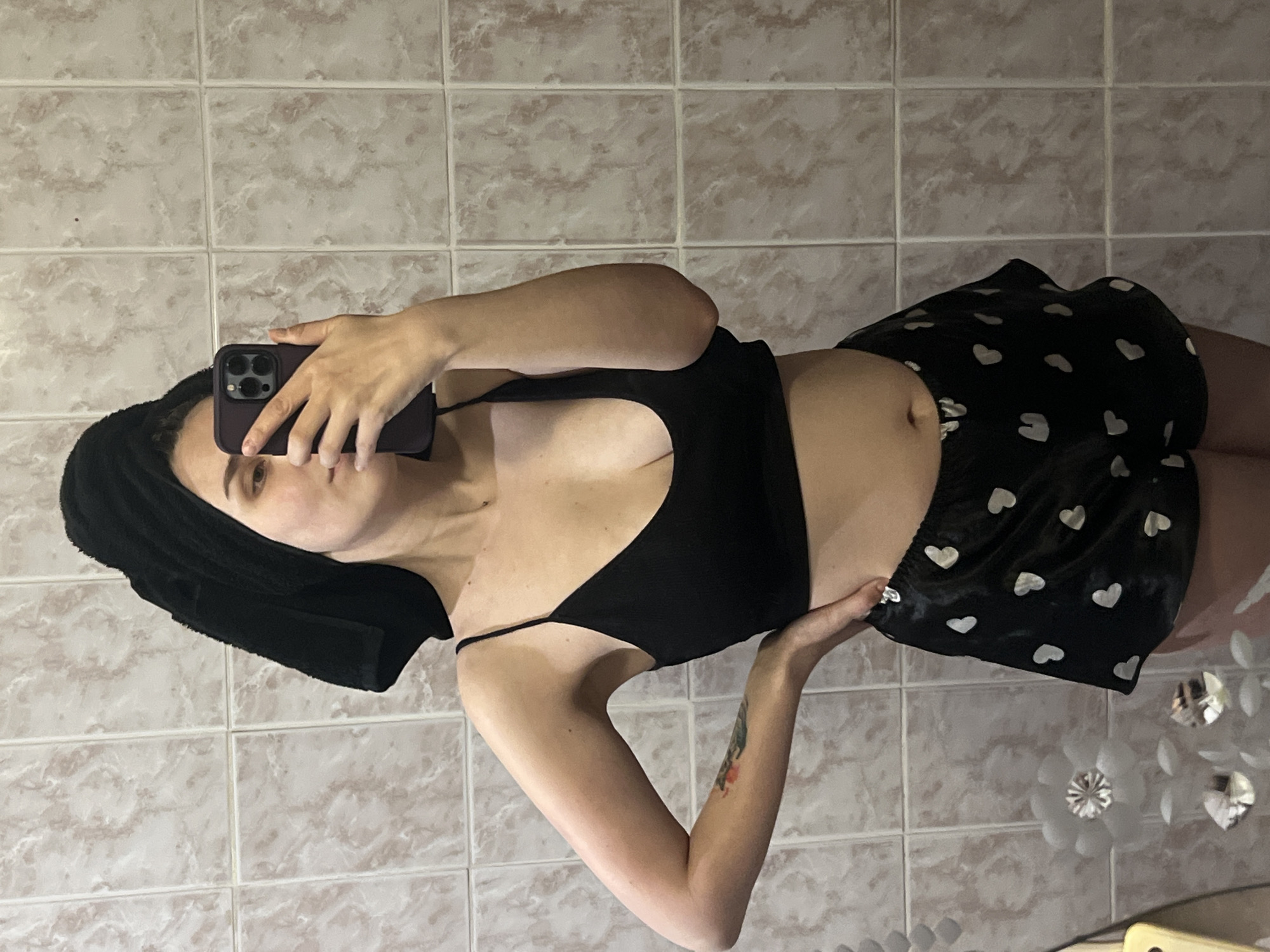 A young woman is depicted taking a mirror selfie in an indoor setting, likely a bathroom or dressing area in Votkinsk, Russia. She stands against a light beige tiled wall featuring a subtle marbled pattern. Her head is wrapped in a black towel or hair wrap, and she is dressed in a black spaghetti strap crop top and black shorts with white heart patterns. She holds a dark-colored smartphone, possibly an iPhone due to the visible camera module, in her right hand, which partially obscures her face. A colorful tattoo is visible on her left forearm. On the right side of the frame, several white, flower-shaped decorative pieces, some appearing reflective, are attached to the tiled wall. The lighting suggests an indoor, artificial light source. The woman's expression is neutral and posed for the self-portrait. No text is visible in the image.