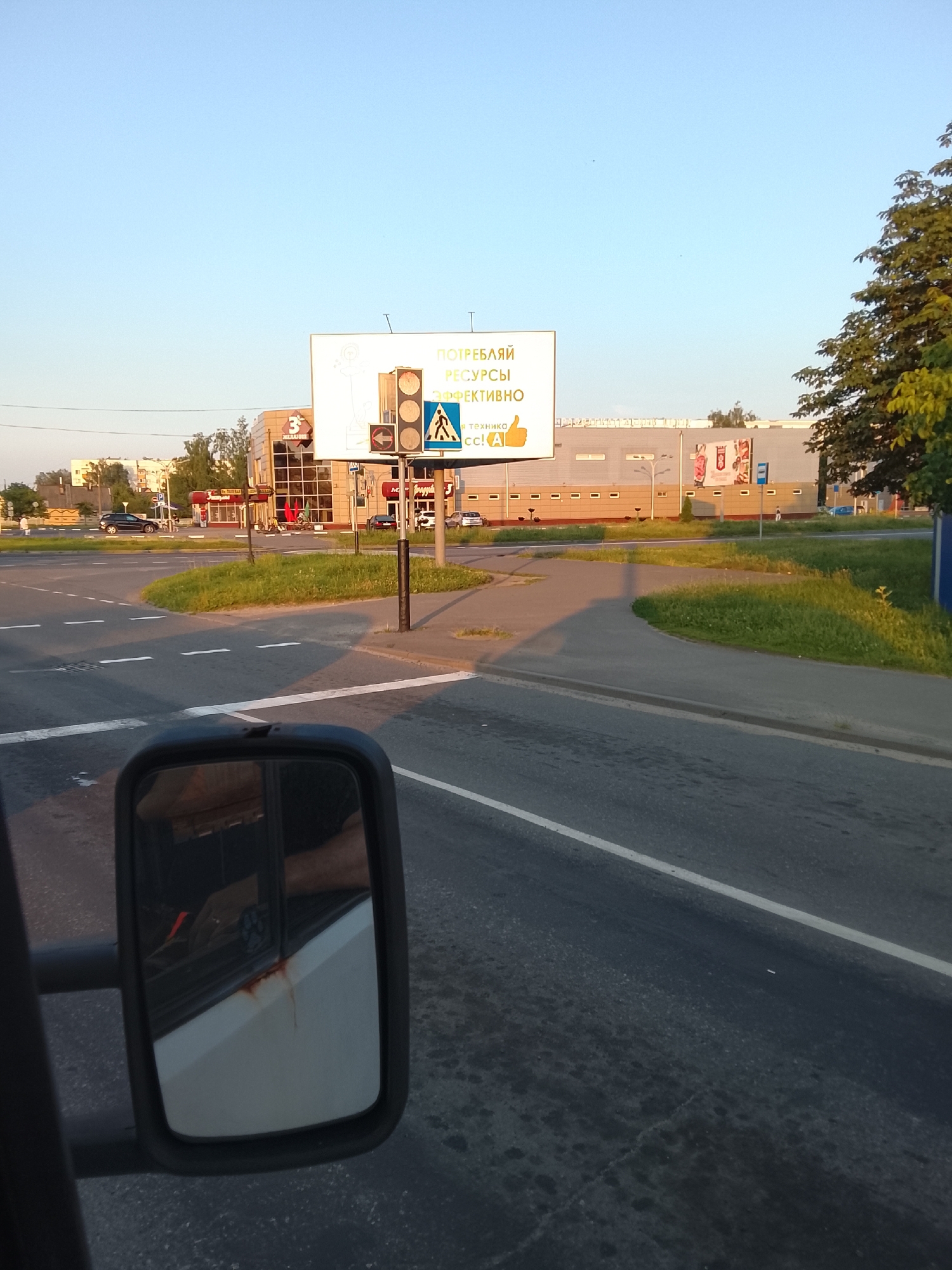 The image captures a street scene in the city of Zhlobin, Belarus, on a clear day with a pale blue sky. In the foreground, a car's side mirror is visible, reflecting the interior of the vehicle and a portion of the passenger's arm. The mirror is mounted on the exterior of the vehicle.
Dominating the midground is a large billboard displaying text in Russian. The billboard's text, which appears to be an advertisement, includes phrases like "ПОТРЕБЛЯЙ РЕСУРСЫ ЭФФЕКТИВНО" (Consume resources efficiently) and "и техника" (and technology). Below the main text, a cartoon thumbs-up icon is present. A traffic light is positioned in front of the billboard, with its signals illuminated. A pedestrian crossing sign is also visible nearby.
Behind the billboard, a modern-looking building with large windows and a beige facade is situated. Several smaller shops with red and brown awnings are also present in the vicinity. A tree with green foliage is seen on the right side of the frame, partially obscuring the building.
The foreground also shows a road with white lane markings and a grassy area with a paved walkway. The overall impression is that of an urban environment with commercial establishments and traffic infrastructure. The lighting suggests it might be late afternoon or early evening due to the long shadows cast by the traffic light pole and the car.
