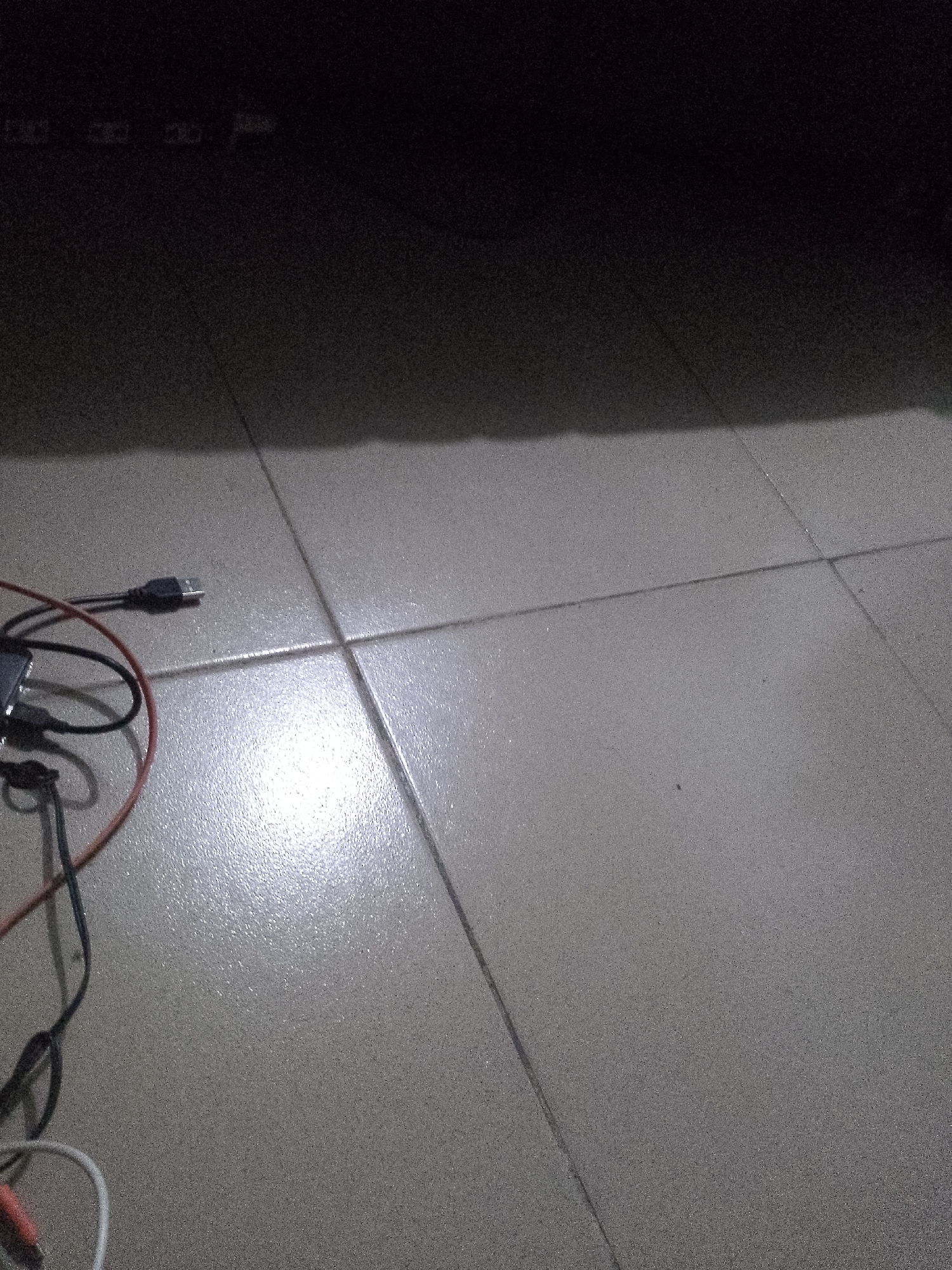 The image shows a close-up view of a tiled floor in Jauro Yinu, Nigeria. Several cables are lying on the floor. One black USB cable is plugged into a black adapter. A red wire and a black wire are also visible, connected to the adapter. A white cable with an orange connector is present at the bottom left of the image. The floor appears to be made of light-colored square tiles, with dark grout lines separating them. A shadow is cast across the top portion of the image, indicating a light source from the upper left. The scene is dimly lit, and the floor has a slight sheen, suggesting it may be slightly damp or reflective.