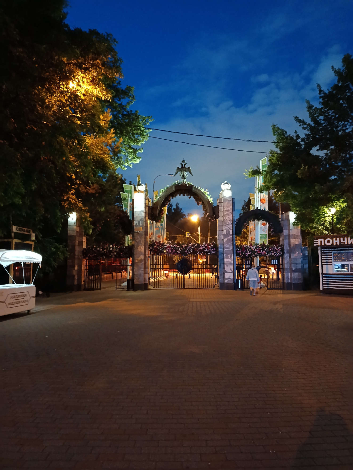 The image captures an illuminated park or venue entrance at night in Moscow, Russia. A grand, ornate archway stands centrally, adorned with garlands of pink and white flowers. Architectural details include a decorative crest, possibly a double-headed eagle, at the apex of the central arch, and elaborate pillars. The sky is a deep blue with scattered clouds, indicating early evening or twilight.
Warm yellow light from streetlights illuminates the dense green foliage of trees flanking both sides of the entrance, as well as the reddish-brown brick-paved ground. One person, seen from behind, is walking towards and through the central gate of the archway.
On the left side of the entrance, a white kiosk or cart is visible, bearing the text "НАПИТКИ МОРЖЕННОЕ," which translates to "Drinks Ice Cream." Above this, a small sign reads "ОХРАНА," meaning "Security." To the right of the archway, a striped kiosk displays the word "ПОНЧИКИ" in large Cyrillic letters, translating to "Donuts." The scene suggests a welcoming, calm atmosphere.