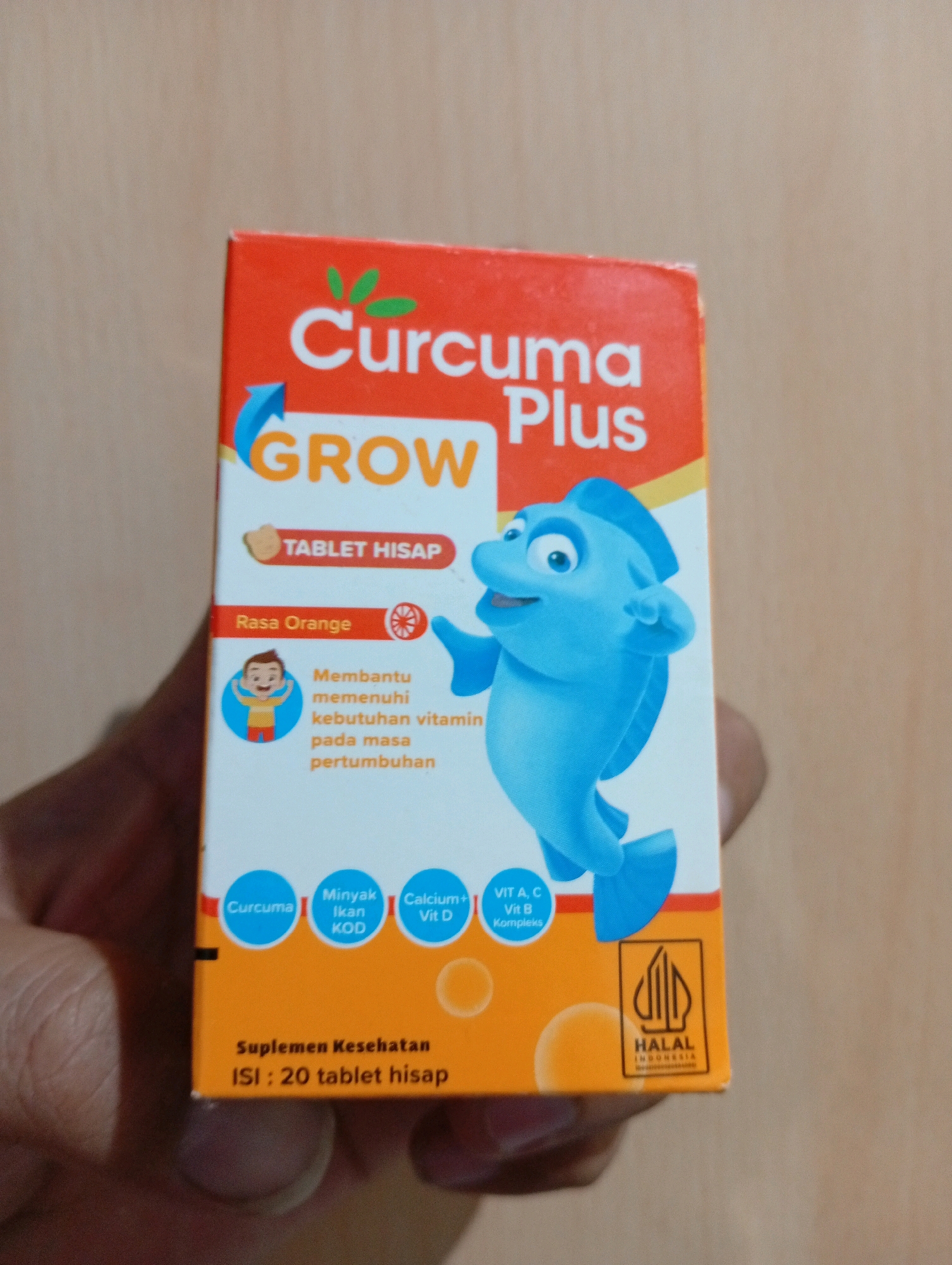 An overhead shot shows a hand holding an orange and yellow box of "Curcuma Plus GROW" supplements. The box features a cartoon blue fish and depicts a child. The text on the box is in Indonesian and indicates that it is a "tablet hisap" (lozenge), flavored orange, and contains Curcuma, fish oil, Calcium, Vitamin A, C, and B complex. The bottom of the box shows "Suplemen Kesehatan" (Health Supplement) and "Isi: 20 tablet hisap" (Contents: 20 lozenges). A Halal certification mark for Indonesia is visible on the right side. The background is a neutral, light brown surface, possibly a table. There is no discernible urban landscape or reference to Kebondalem, Indonesia, in the image.