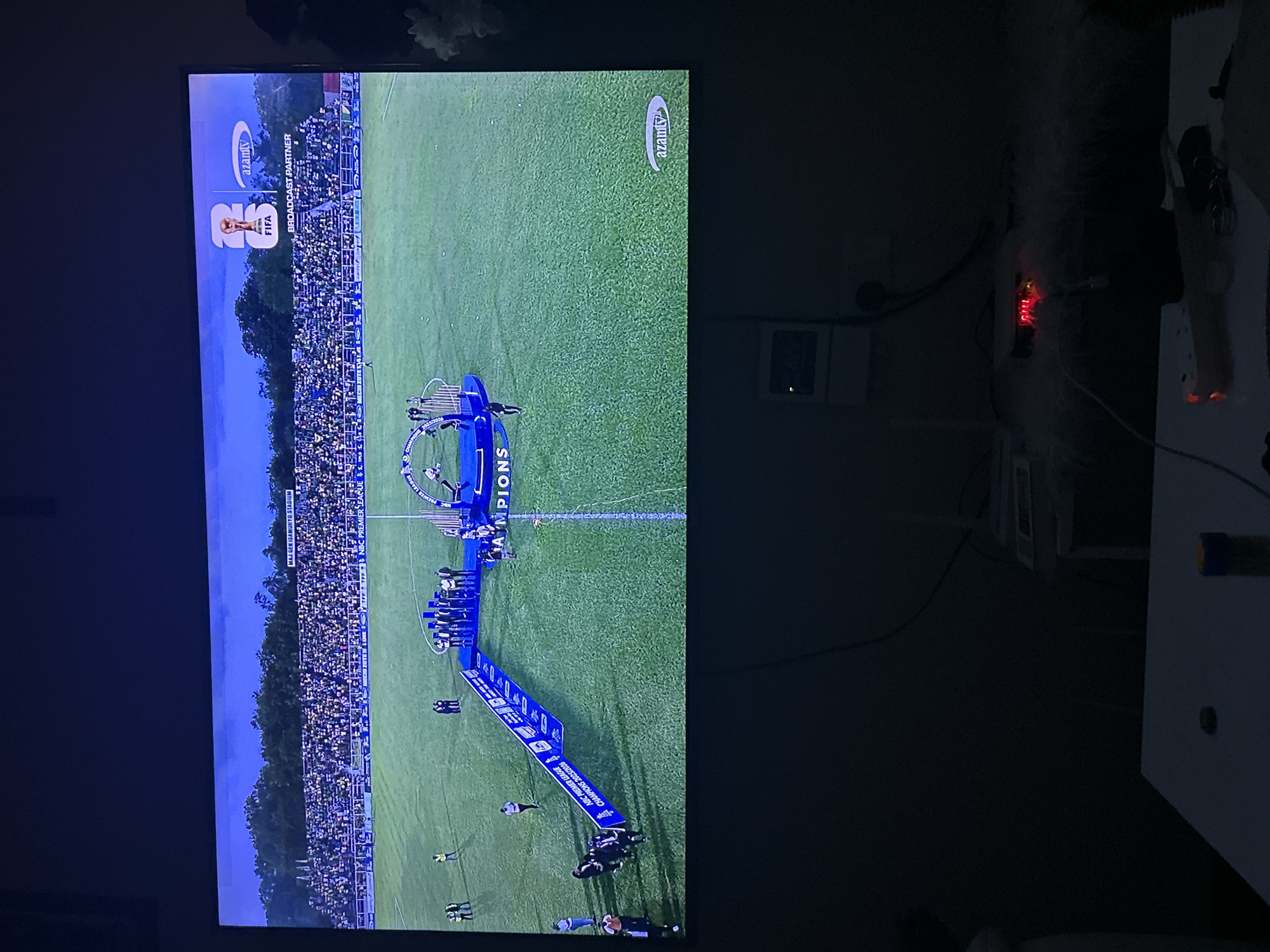This media file depicts a nighttime scene on a soccer field, likely in Dar es Salaam, Tanzania, as suggested by the "NBC PREMIER LEAGUE" text. A trophy presentation ceremony is underway.
The main subjects are a group of people, presumably soccer players and staff, gathered on the field. Some are standing on a blue platform or stage, while others are on the grass. They appear to be celebrating, as indicated by the joyful atmosphere of a trophy presentation.
Several objects are visible. There's a large blue banner or backdrop on the field that reads "NBC PREMIER LEAGUE CHAMPIONS 2023/2024". There are also what appear to be trophy stands or risers, and possibly a trophy itself is present on the field, though not clearly visible. The field is covered in green grass. In the background, stadium seating filled with spectators can be seen. The sky is dark, indicating it is nighttime.
The activity taking place is the celebration and trophy presentation for the NBC Premier League champions. The emotion conveyed is one of triumph and celebration.
Visible text includes "NBC PREMIER LEAGUE", "CHAMPIONS 2023/2024", "FIFA", and "AZAMTV". The latter two are likely logos of broadcast partners or official organizations. The text "MAJ G Gen GARIBOTO STADIUM" is also visible on a sign in the background, providing a specific location cue.