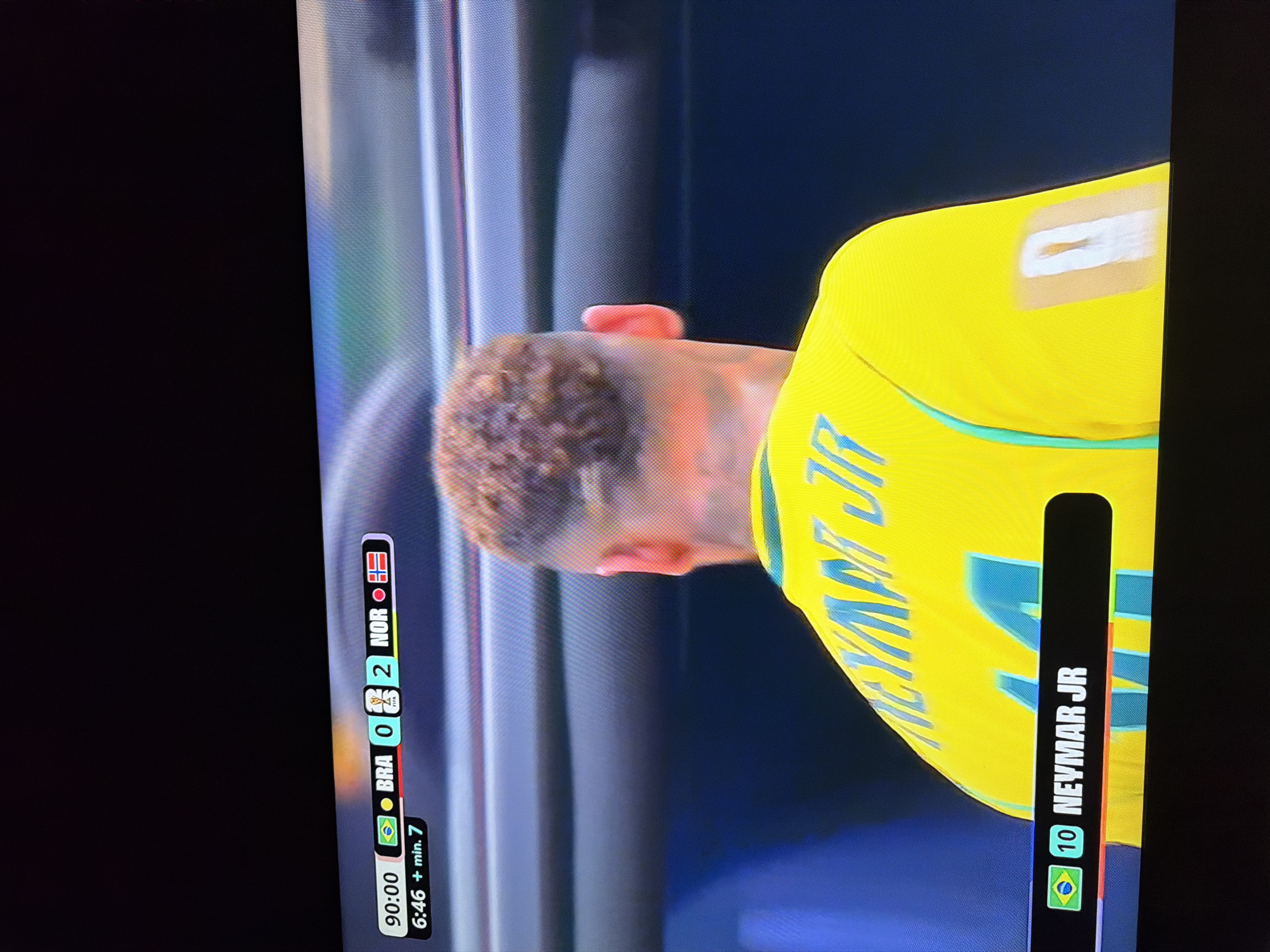 The image displays a television screen showing a soccer match scoreboard and a player's back. The scoreboard indicates the time is 90:00 and the score is BRA 0 - 2 NOR. Below that, it shows 6:46 + min. 7. The player in the foreground is wearing a yellow jersey with "NEYMAR JR" and the number "10" visible on the lower left of the screen, next to a Brazilian flag. A small bandage is visible on the player's upper right arm. The background of the image shows a stadium seating area. The image quality suggests it was taken in a dimly lit room, likely a home or a bar, with the television screen being the primary light source. There is no information to suggest this scene is in Ognianovo, Bulgaria.