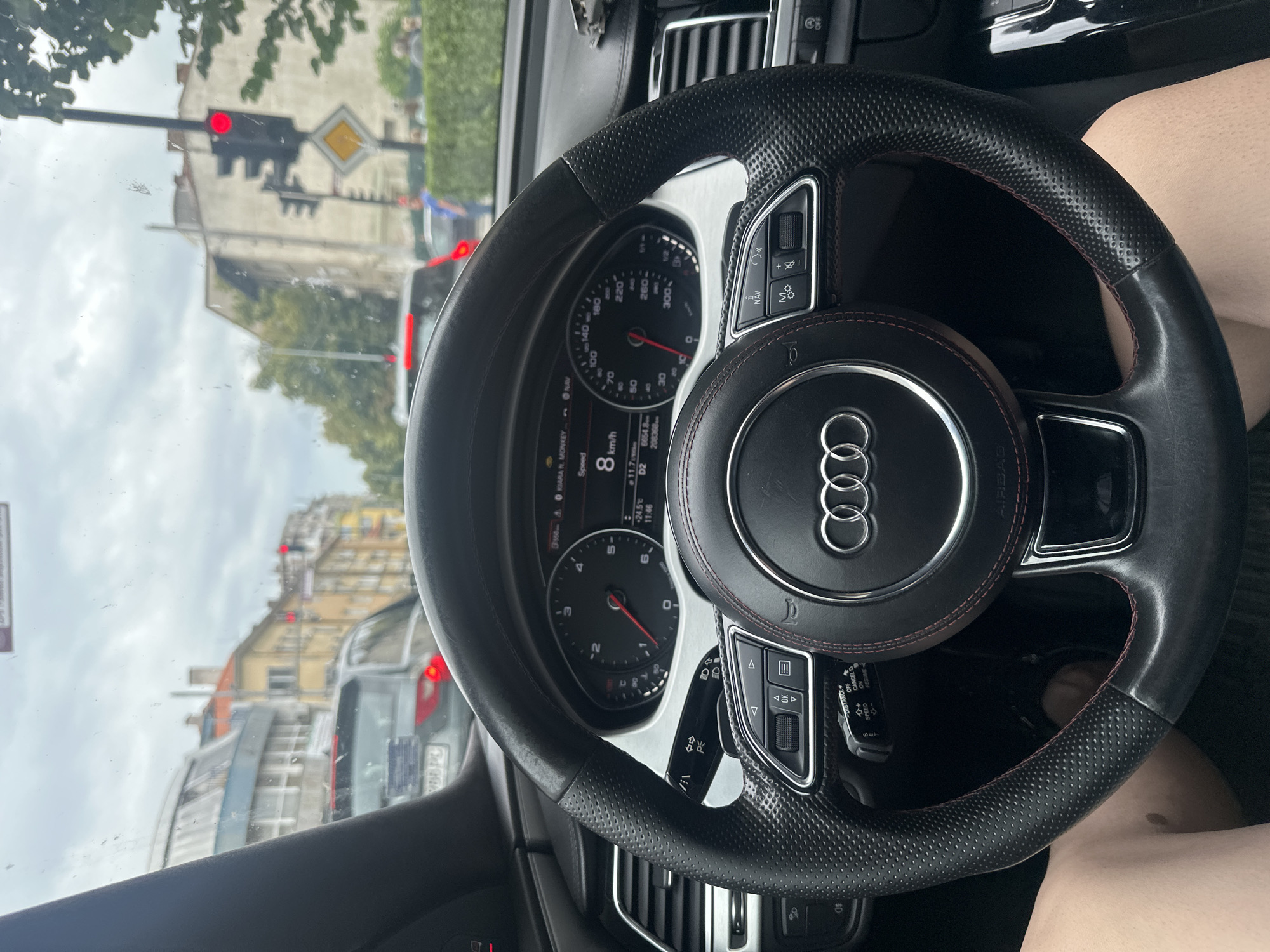 The image is a first-person perspective from inside a car, looking at the steering wheel and dashboard. The car is in traffic, evidenced by the red traffic light ahead and other vehicles. The dashboard displays a speed of 8 km/h. The exterior view shows buildings lining a street, with trees and a cloudy sky. The location is identified as Kazanlak, Bulgaria.