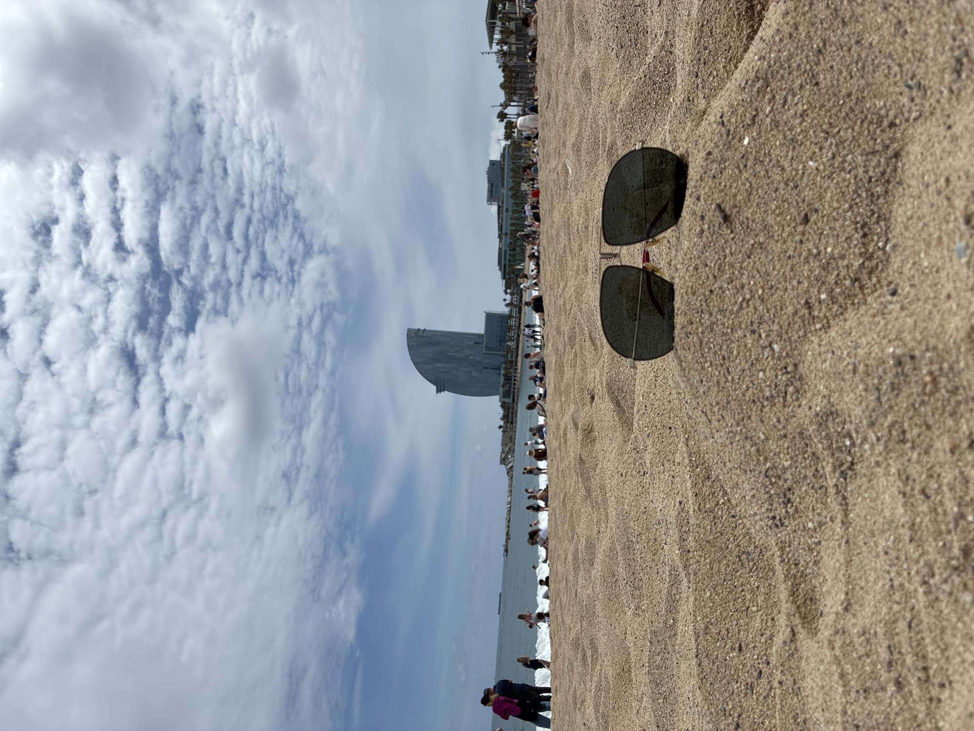 The image depicts a beach scene, likely in Barcelona, Spain. A pair of sunglasses rests on the sand in the foreground. In the background, a prominent building resembling a sail, known as the W Barcelona Hotel, is visible. People are seen relaxing on the beach and near the water's edge. The sky is partly cloudy, with formations covering a significant portion of the frame.