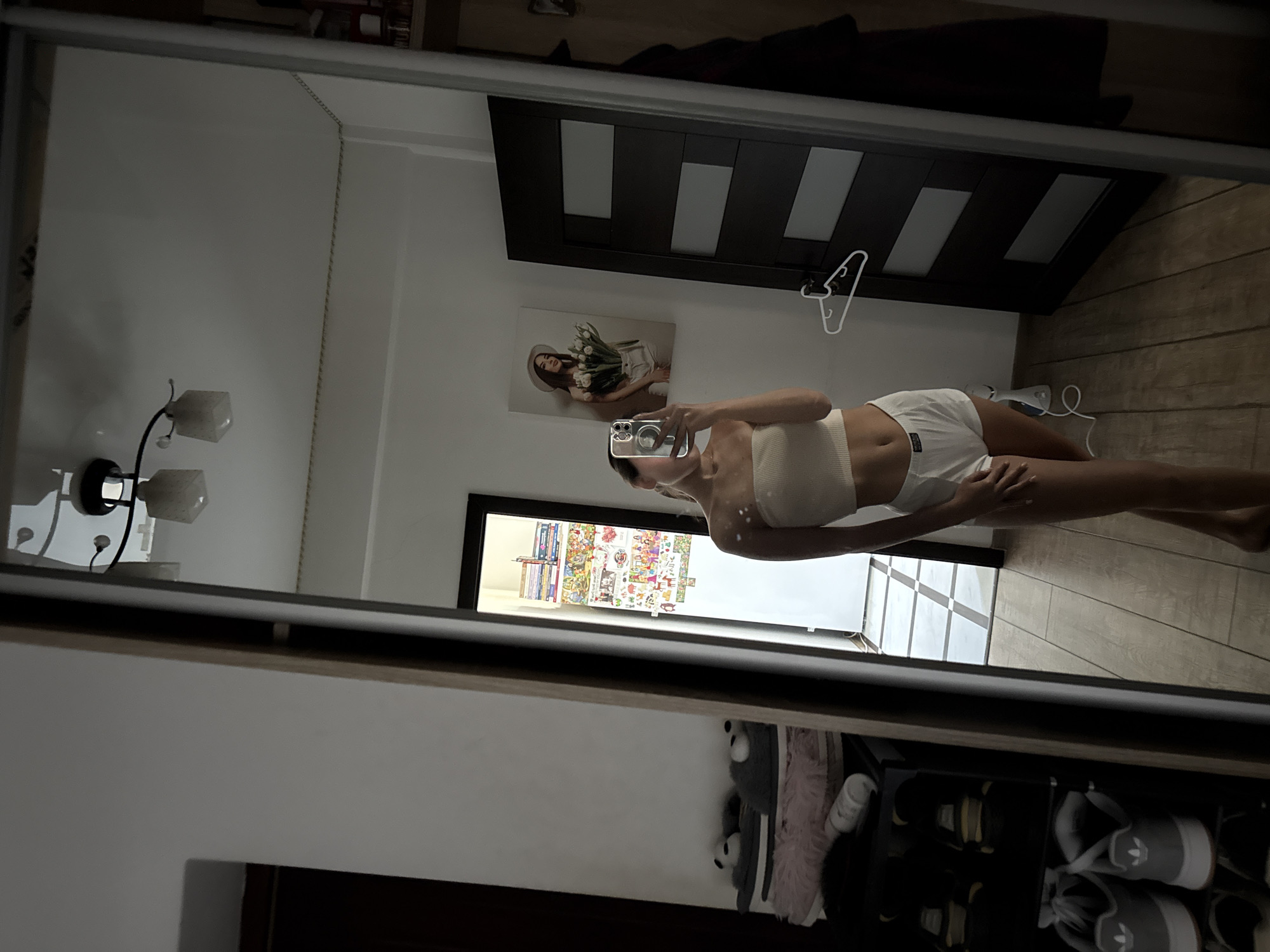 A young woman, seen from the waist down, is taking a mirror selfie in what appears to be a bedroom. She is wearing a white tube top and white shorts. The mirror is large and framed, reflecting the room behind her. To her left, a closet with shoes and stuffed animals is visible. To her right, a dark brown door with frosted glass panels is present. On the wall behind her, a framed picture of a woman holding flowers and a hat is displayed. The floor is made of light-colored wood planks. The overall impression is that of a casual, domestic setting. There is no indication that this location is in Chișinău, Moldova.