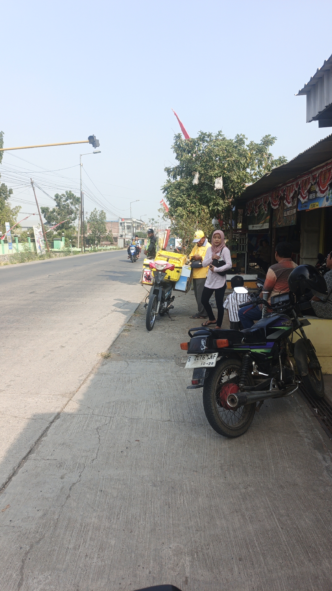 The image depicts a street scene in Sumengko, Indonesia, likely on a sunny day with a clear blue sky. The foreground is dominated by a concrete sidewalk and road. Several motorcycles are parked on the sidewalk. One prominent motorcycle in the mid-ground has a bright yellow cargo box attached to its rear.
Two individuals are standing near this motorcycle. One is a woman wearing a pink headscarf and a striped shirt, holding a black bag. The other is a man wearing a yellow cap and a yellow shirt, also holding a bag. They appear to be interacting with the motorcycle or its cargo.
Behind them, a building with a corrugated metal roof and a striped awning stretches along the right side of the image. Several people are visible inside or in front of this building, seated and seemingly engaged in activity, though their specific actions are not clear. Another motorcycle is parked to the right of the building.
In the background, a street with traffic is visible. A few more motorcycles are on the road, and some buildings and trees line the street. Traffic signals and power lines are also present, indicating an urban or semi-urban environment. Indonesian flags are visible in the distance, adding a cultural context to the scene. The overall impression is one of a busy, everyday scene in a roadside commercial area.