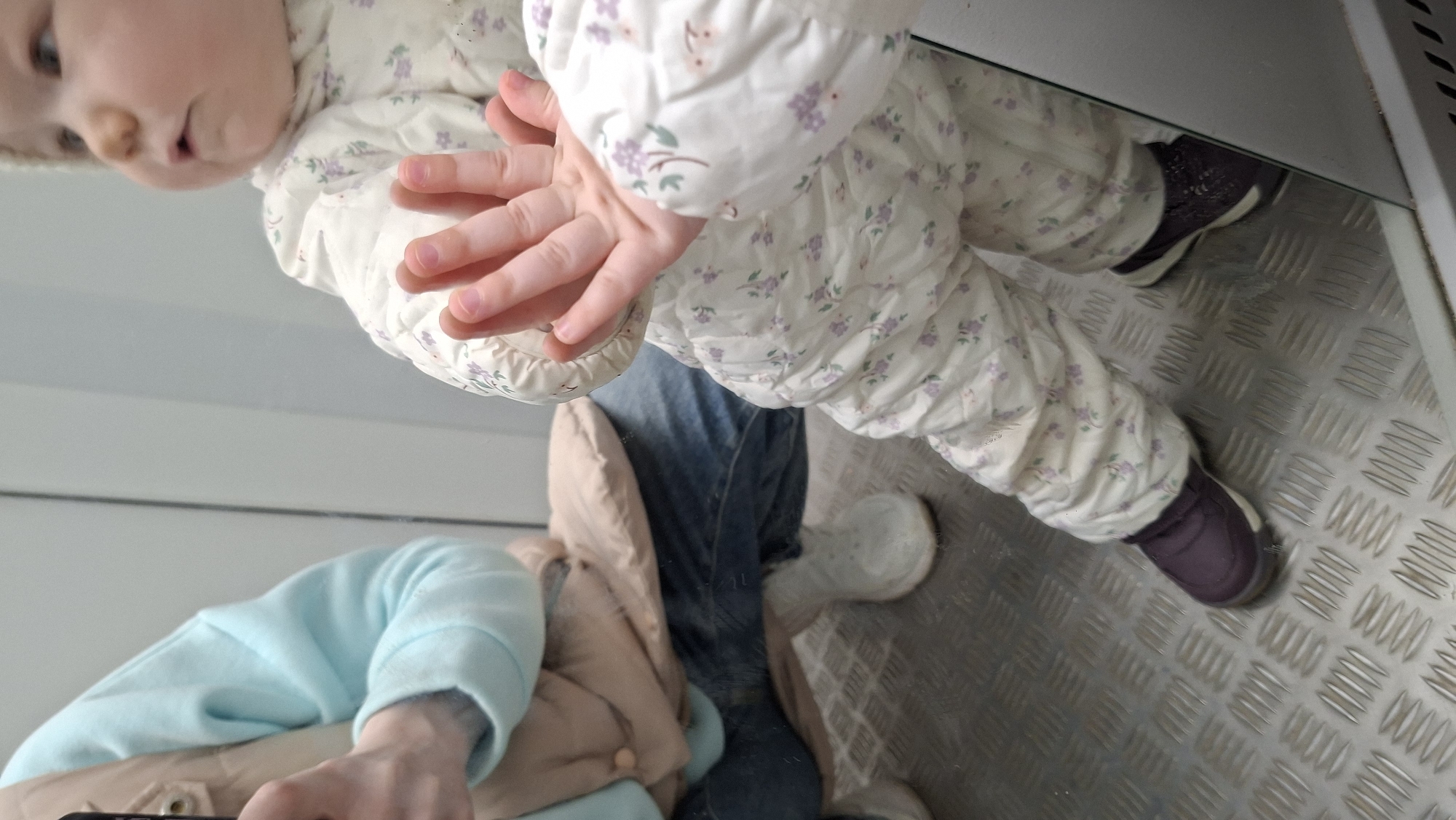 A reflection image depicts a young child in a floral-patterned winter coat and pants, along with a person dressed in blue jeans and a light blue shirt under a beige jacket. The child is positioned towards the top-left of the frame, with their hands near the center. The person's arm and jacket are visible in the bottom-left. The reflection is captured on a diamond-plated metal surface, with a mirror edge running along the top. The location is an indoor area in a city of Unknown, Unknown.