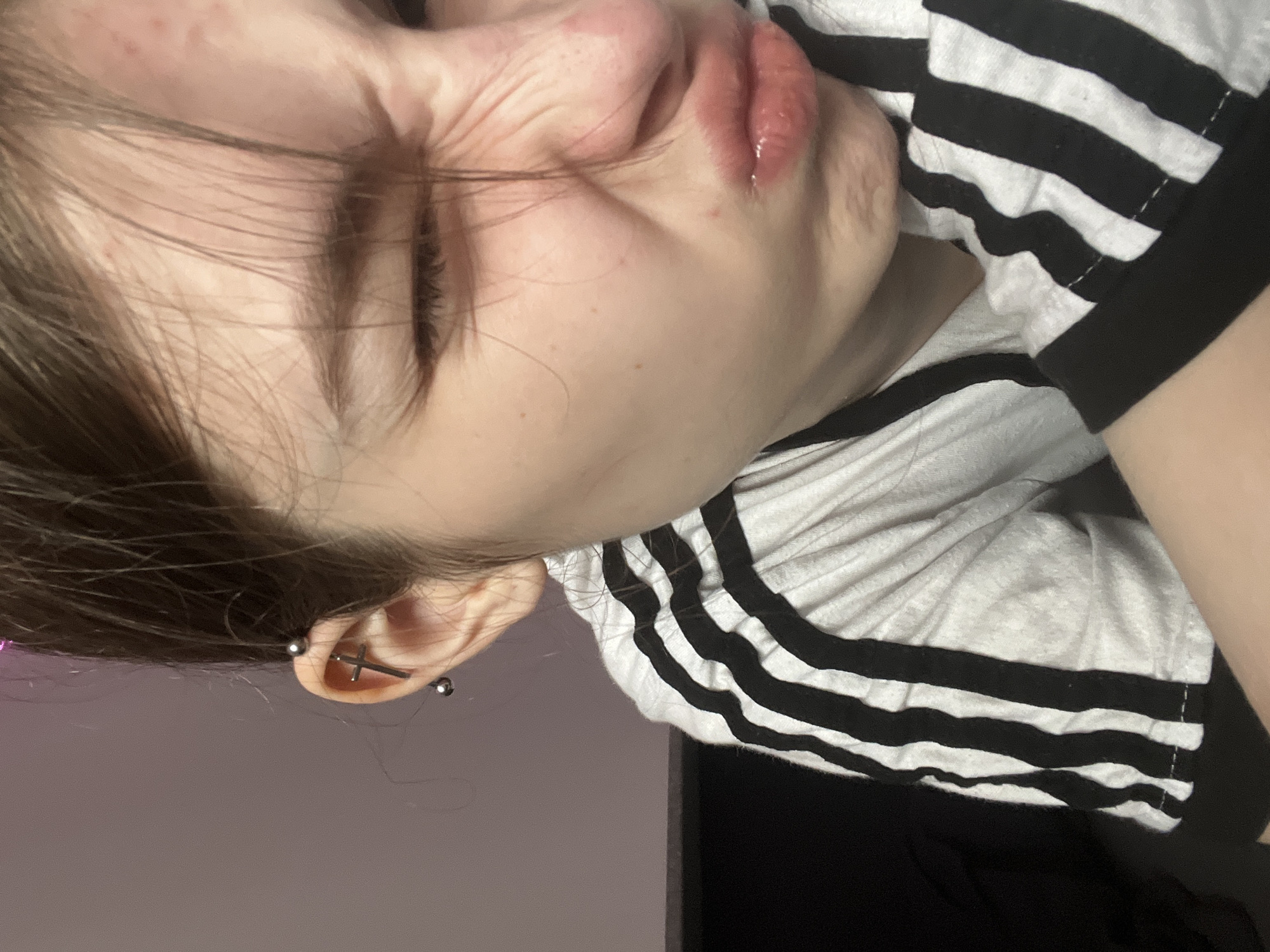 The image displays a close-up, rotated portrait of a young individual, possibly making a playful or exaggerated facial expression. The person's head is tilted to the side, with brown hair partially obscuring the forehead. Their eyes are squinted shut, creating prominent wrinkles around the eye area and bridge of the nose. The lips are pursed and appear slightly glossy or wet. Scattered small red blemishes are visible on the forehead and cheek area.
The individual is wearing a white t-shirt with bold black horizontal stripes on the sleeve, reminiscent of an athletic apparel brand design. On the visible left ear, there are two distinct piercings: an industrial piercing featuring a silver bar across the upper cartilage, and a standard lobe piercing with a silver cross-shaped earring.
The background is simple and out of focus, appearing to be an indoor setting with a light-colored wall, subtly illuminated with a soft purplish-pink light on the left side, transitioning to a darker area on the right. No visible text is present in the image. While a location context of Saint Petersburg, Russia, was provided, no visual cues within the image itself confirm this setting.
