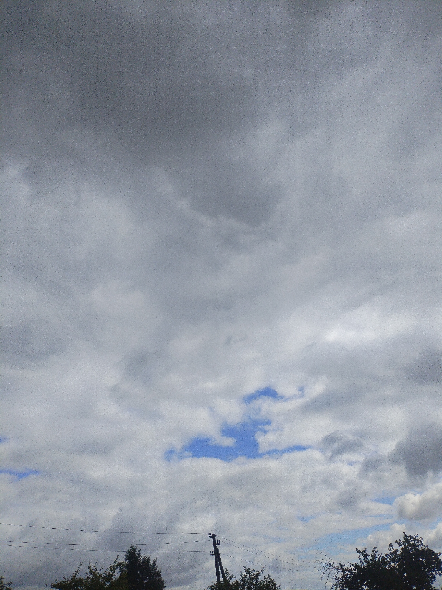 The image shows a cloudy sky above a treeline with utility poles and wires. The sky is predominantly grey with patches of lighter clouds and some glimpses of blue sky. There are no people visible in the image. The foreground features the tops of dark green trees, some bare branches, and two utility poles with attached wires running horizontally across the frame. The scene appears to be outdoors in a rural or suburban setting, consistent with the location of Вароніцкі сельскі Савет, Belarus.
